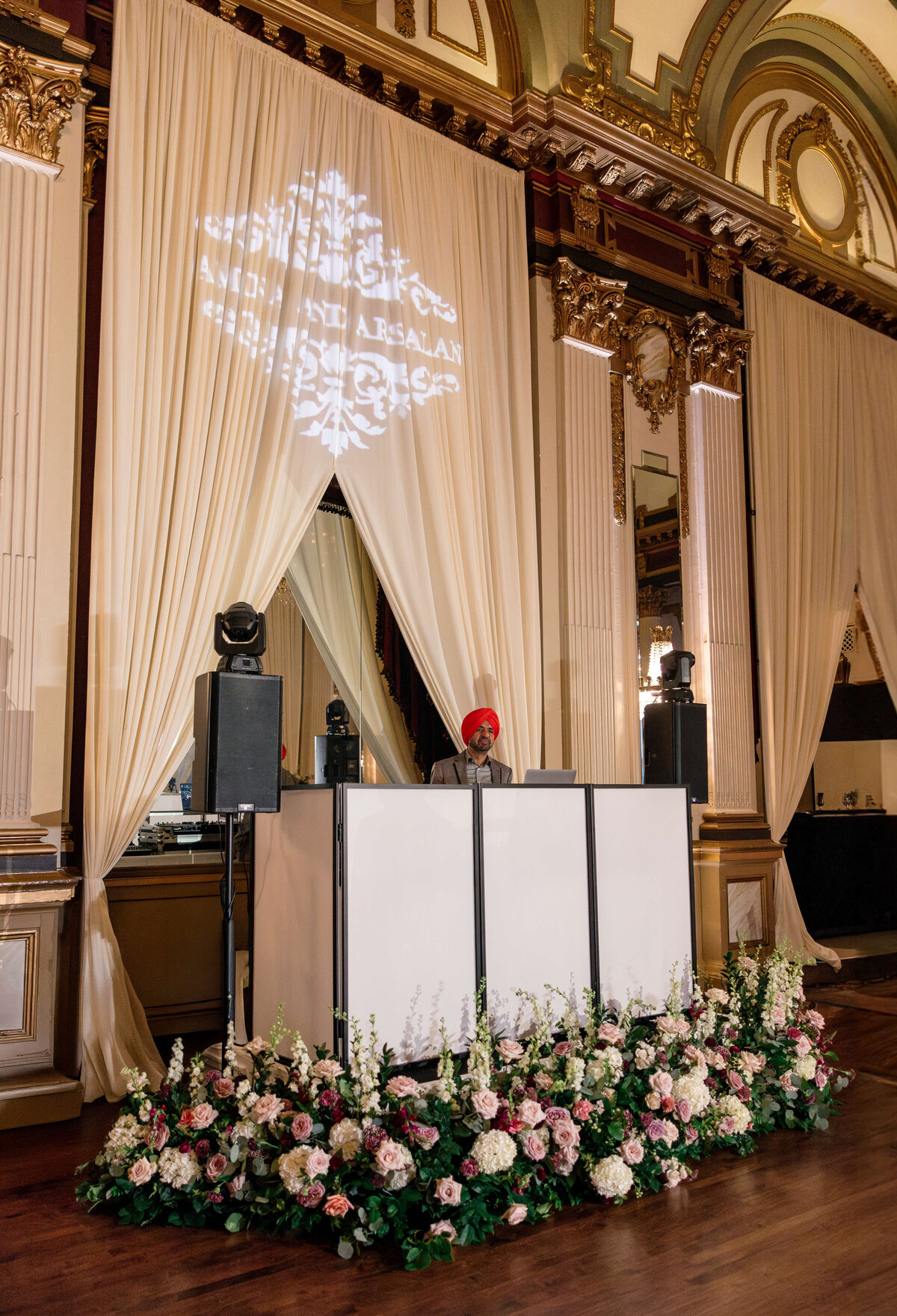 A DJ wearing a bright red turban performs at an elaborate venue with ornate architecture. Behind the DJ booth, white drapes hang, and a floral arrangement adorns the area. A monogram and names are projected on the drapes above.