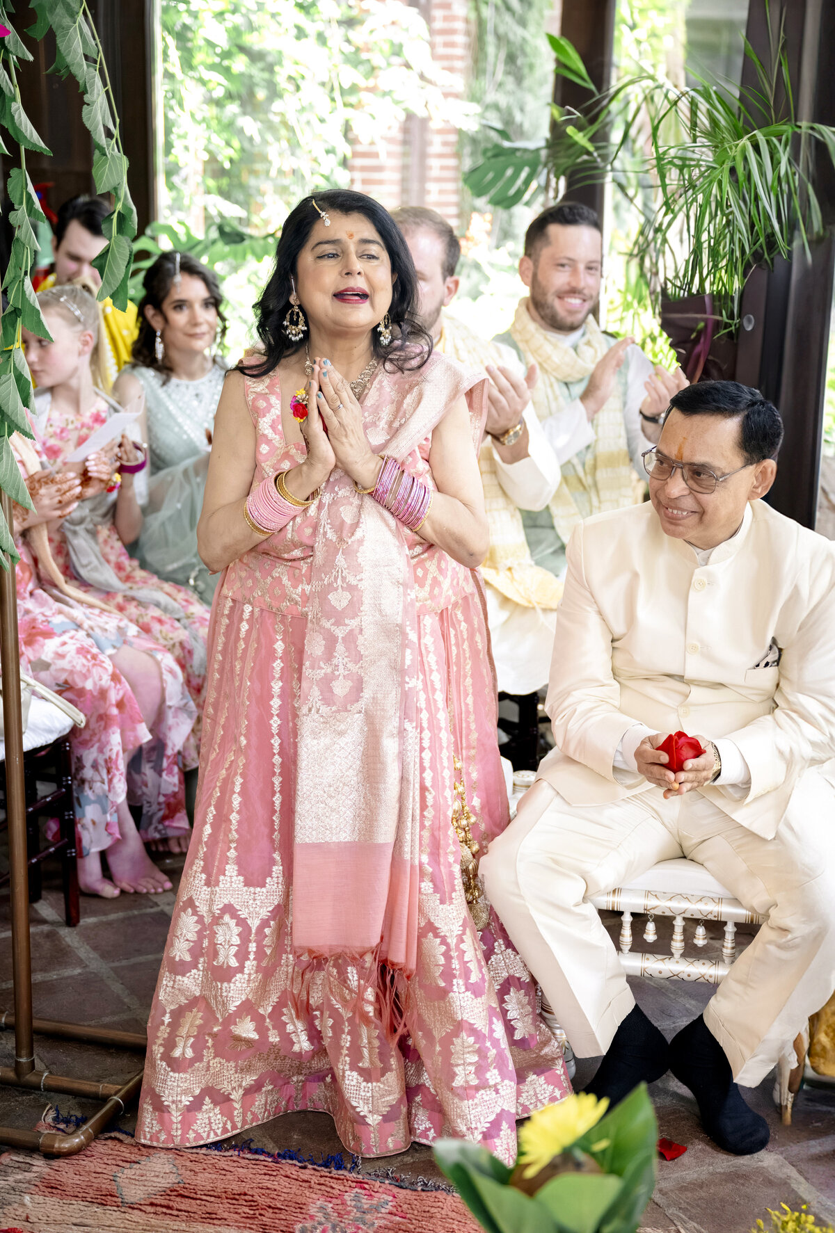 A woman in a pink traditional dress clasps her hands joyfully, standing beside a seated man in a white outfit holding a red flower. They are surrounded by smiling people in colorful attire, set in a bright, plant-filled room.