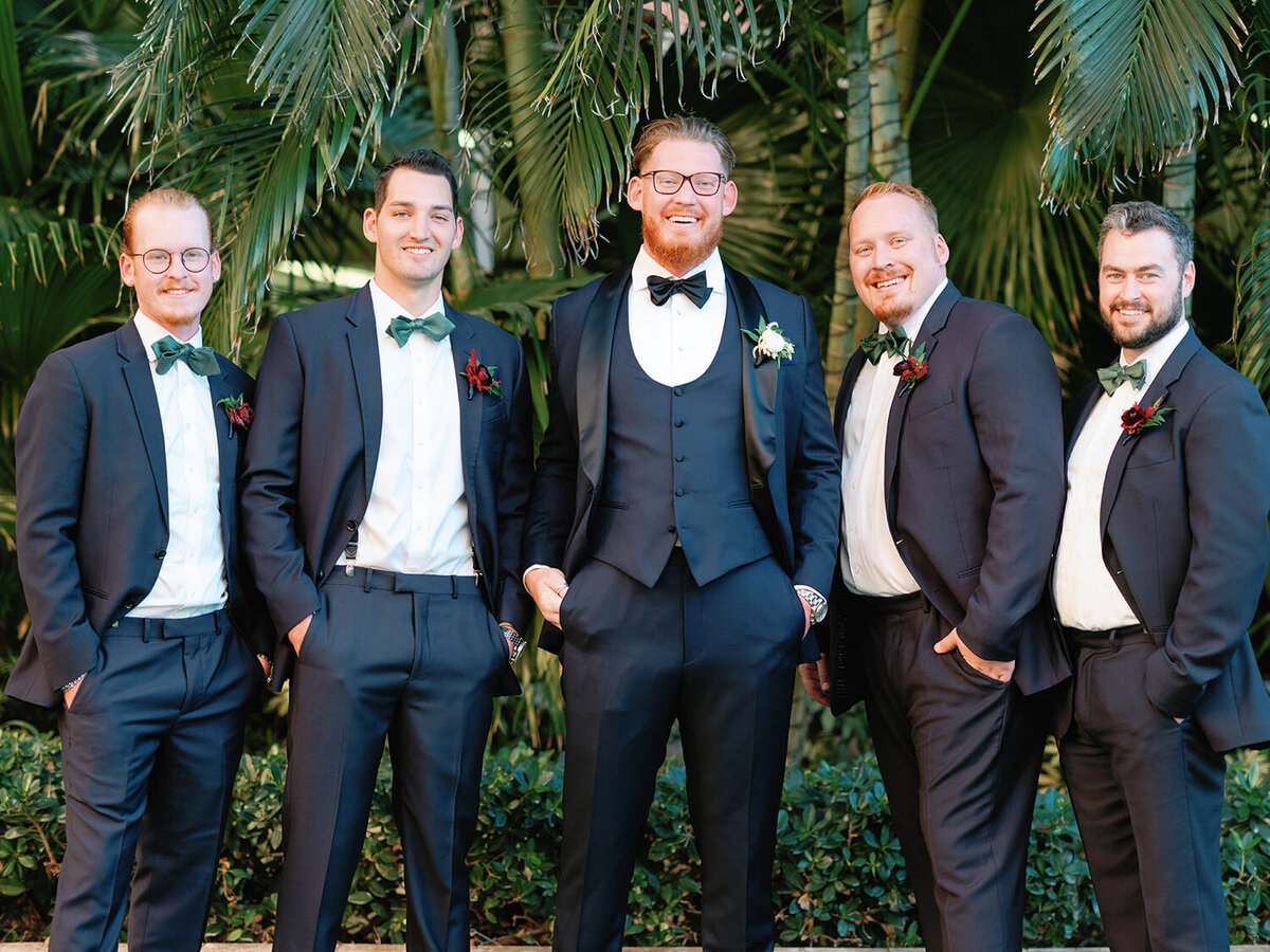 Groom and groomsmen in navy blue tuxedos with emerald green bowties posing against a backdrop of palm trees at the Don Cesar Hotel, St. Pete Beach, FL, for a December wedding.