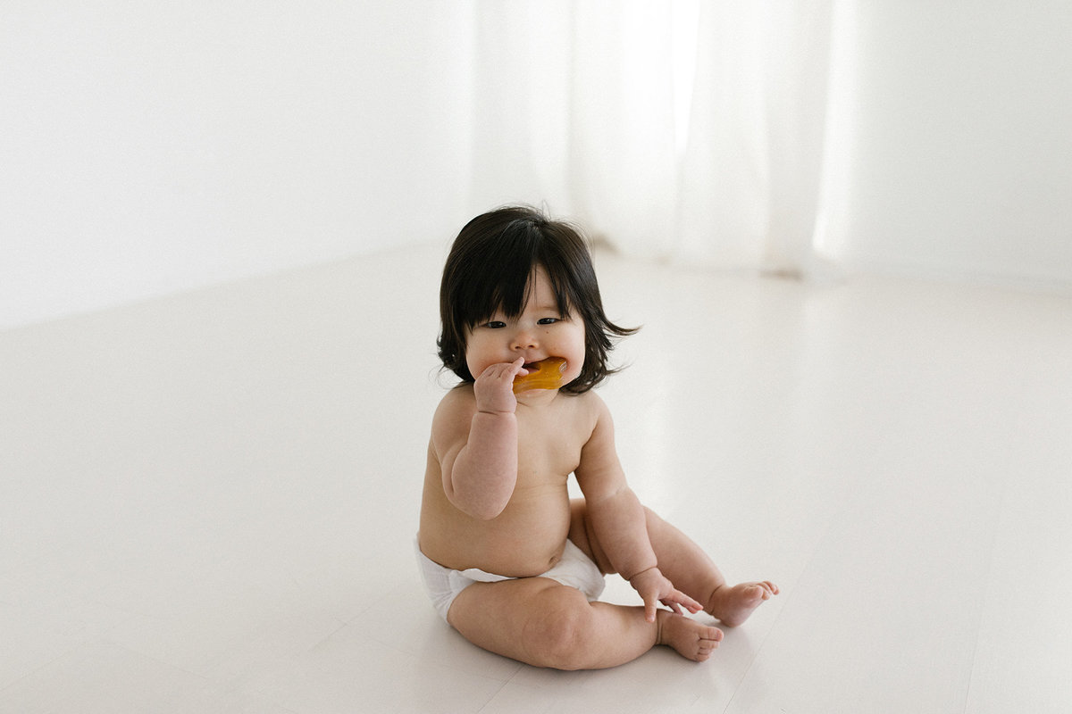 Little baby with dark hair chewing on a teether toy in an all white natural light studio