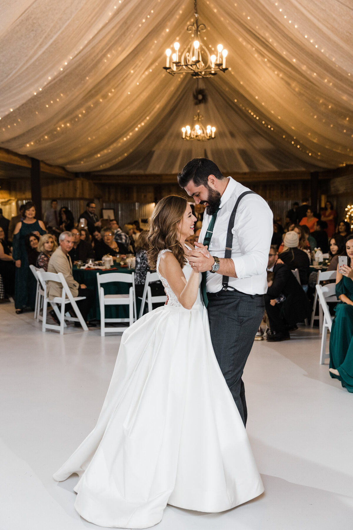 Candid shot of a couple sharing their first dance during their wedding reception in Abilene, Texas. The bride is on the left and is wearing a sleeveless, white dress. The groom is on the left and is wearing a suit (minus the jacket) with a green tie and suspenders. Chandeliers, strings of lights, and flowing draperies hang overhead while many guests can sit in the background at their reception tables.