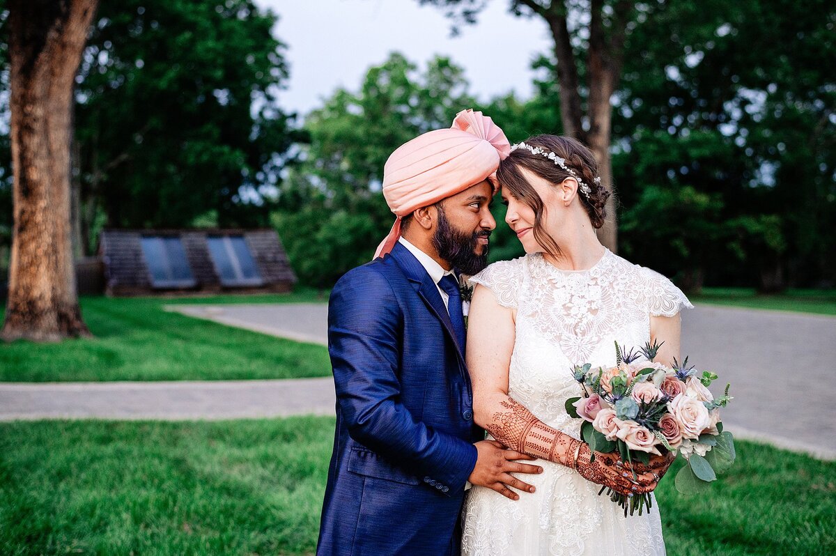 A hindu groom and Christian bride lean in for an intimate moment. The groom is wearing a cobalt blue suit with a blush turban. The bride is wearing a lace wedding dress with cap sleeves and is holding a bouquet of ivory and blush roses accented with crimson dahlias.