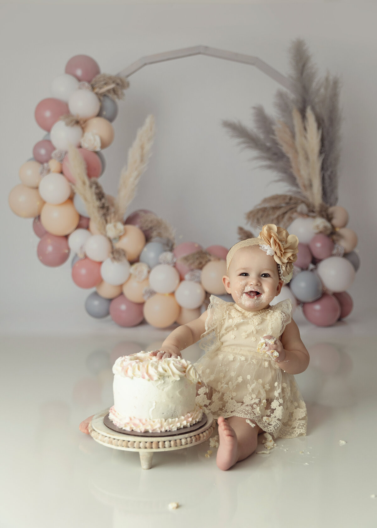 A toddler girl in a cream lace dress smiles while eating a white cake with a balloon backdrop