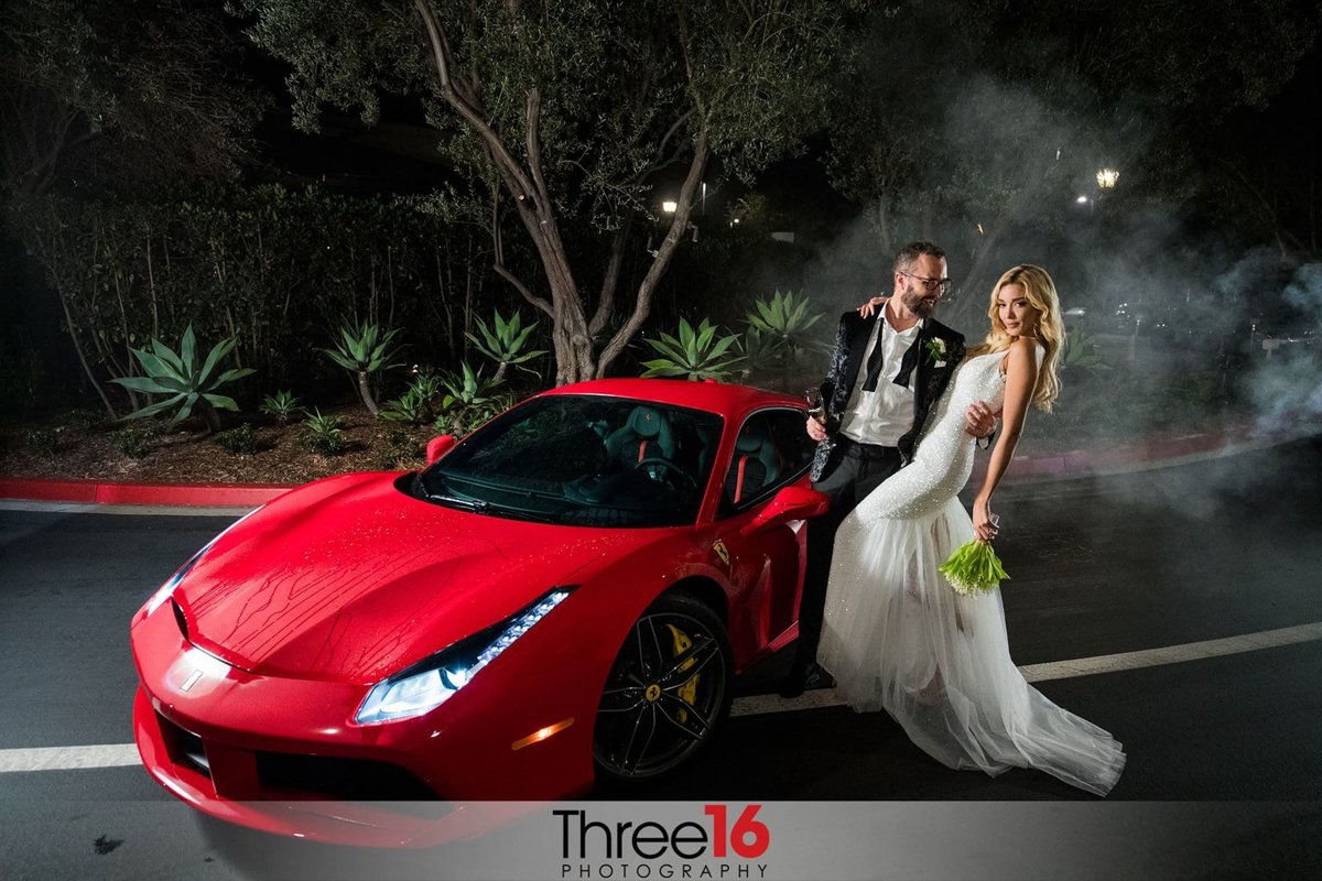 Bride and Groom pose next to a red sportscar with her bouquet down by her side and a glass in his hand