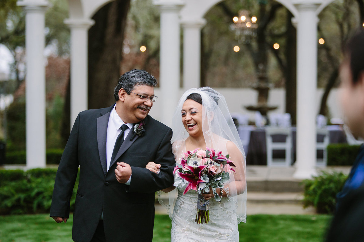 bride and father of bride laugh as walking down aisle during ceremony at Gardens at West Green Venue in San Antonio