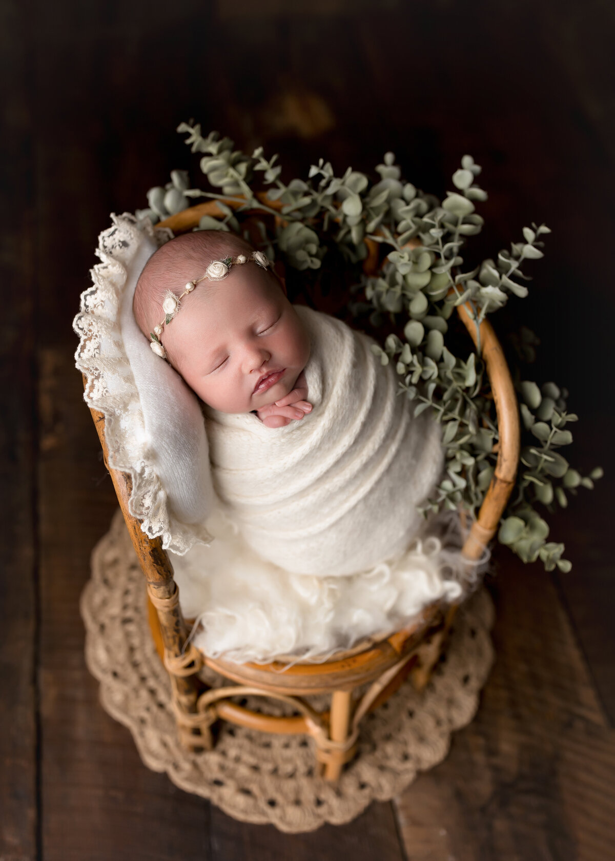 Baby girl sleeping in wicker chair for newborn photoshoot. Baby is wrapped in a cream knit swaddle with a delicate floral headband. Her head is resting on a cream colored lace pillow. Eucalyptus is woven in the chair.