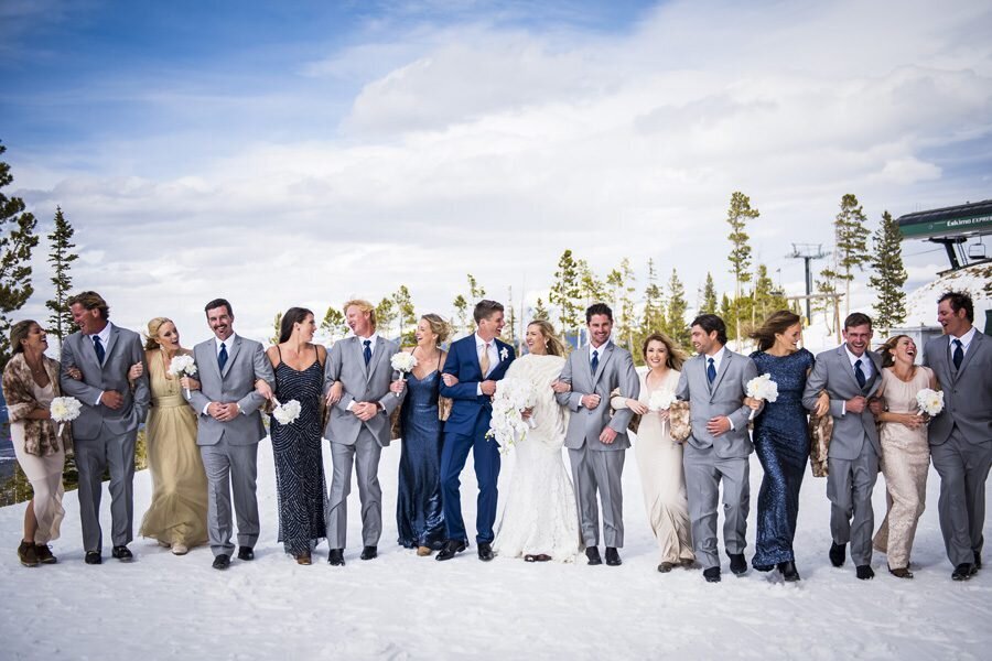 A large wedding party walks toward the camera arm in arm through the snow with the bride and groom in the middle.