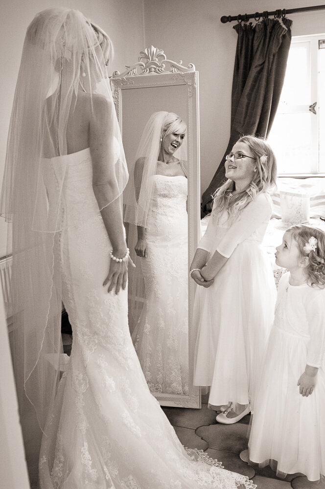 bride with blonde hair wearing veil and trumpet style, sleeveless, lace wedding dress, in front of mirror, smiling at her flowergirls