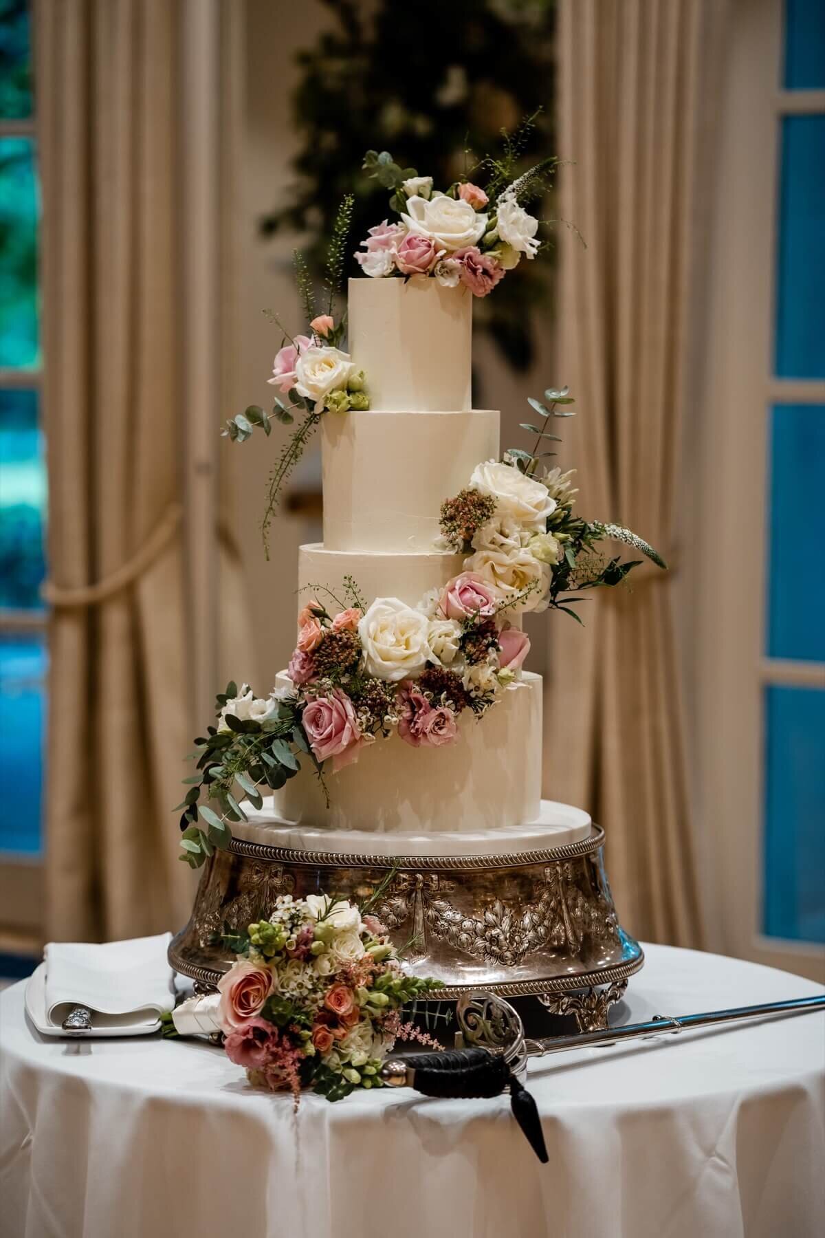 An elegant wedding cake with florals running diagonally down the cake on a silver cake stand with a sword sitting on the cake table