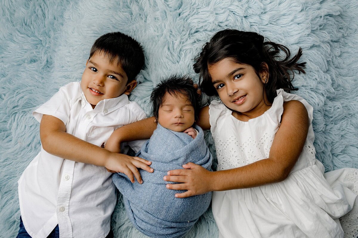 brother and sister dressed in white cuddle newborn baby brother wrapped in blue while laying on a blue rug
