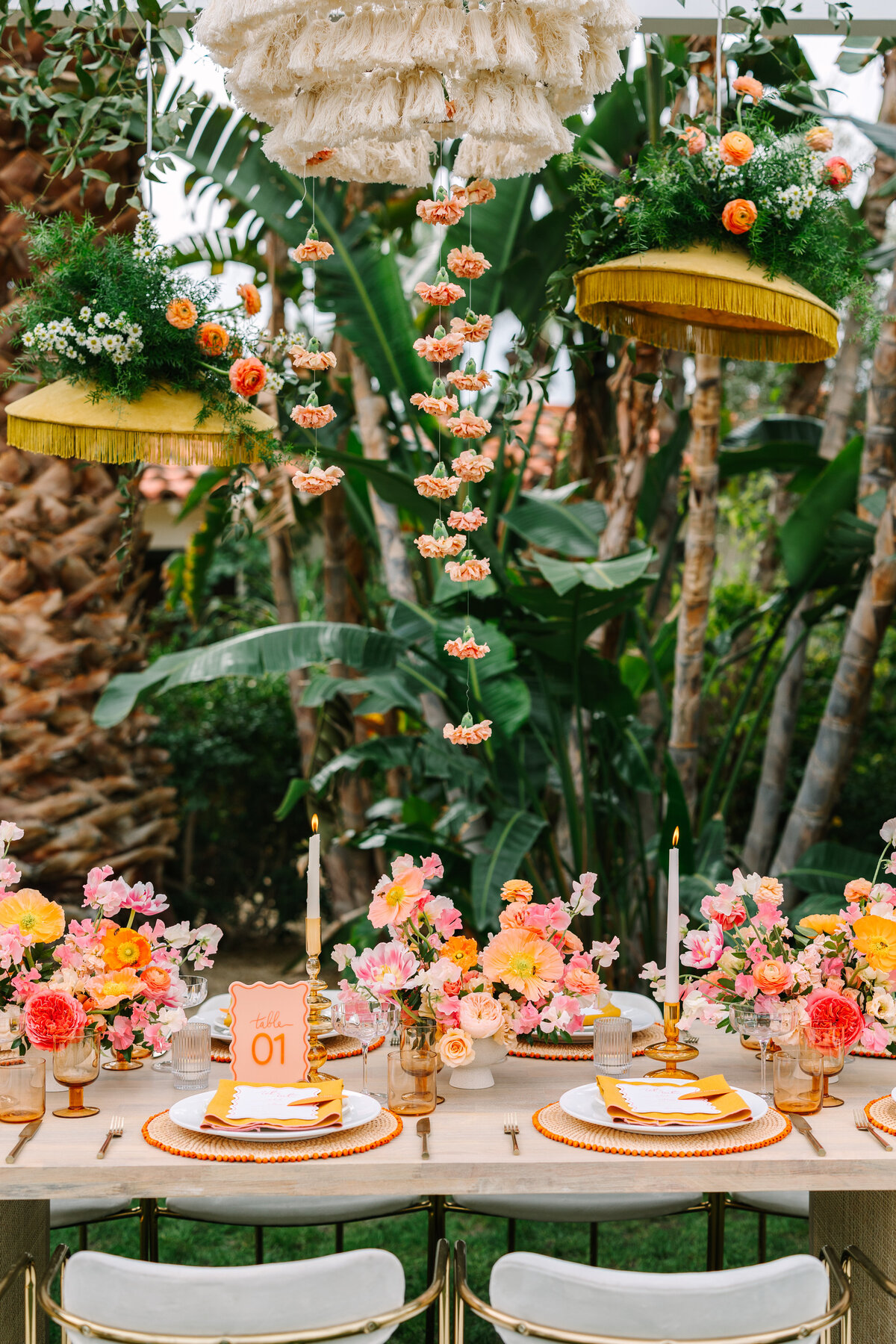 fun and colorful wedding tablescape with orange, pink, and red florals and boho light fixtures with palm trees in background.