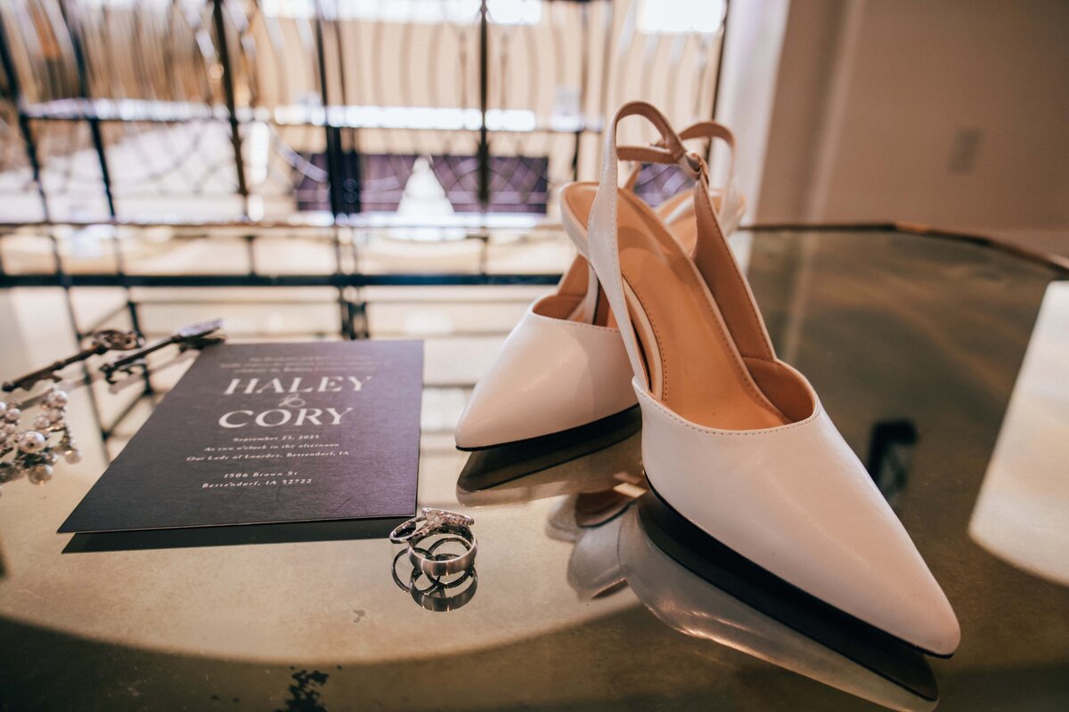 Pair of white high-heeled shoes on a glass table beside a wedding invitation reading "Haley &amp; Cory, March 18, 2023," arranged by a wedding planner in Des Moines.