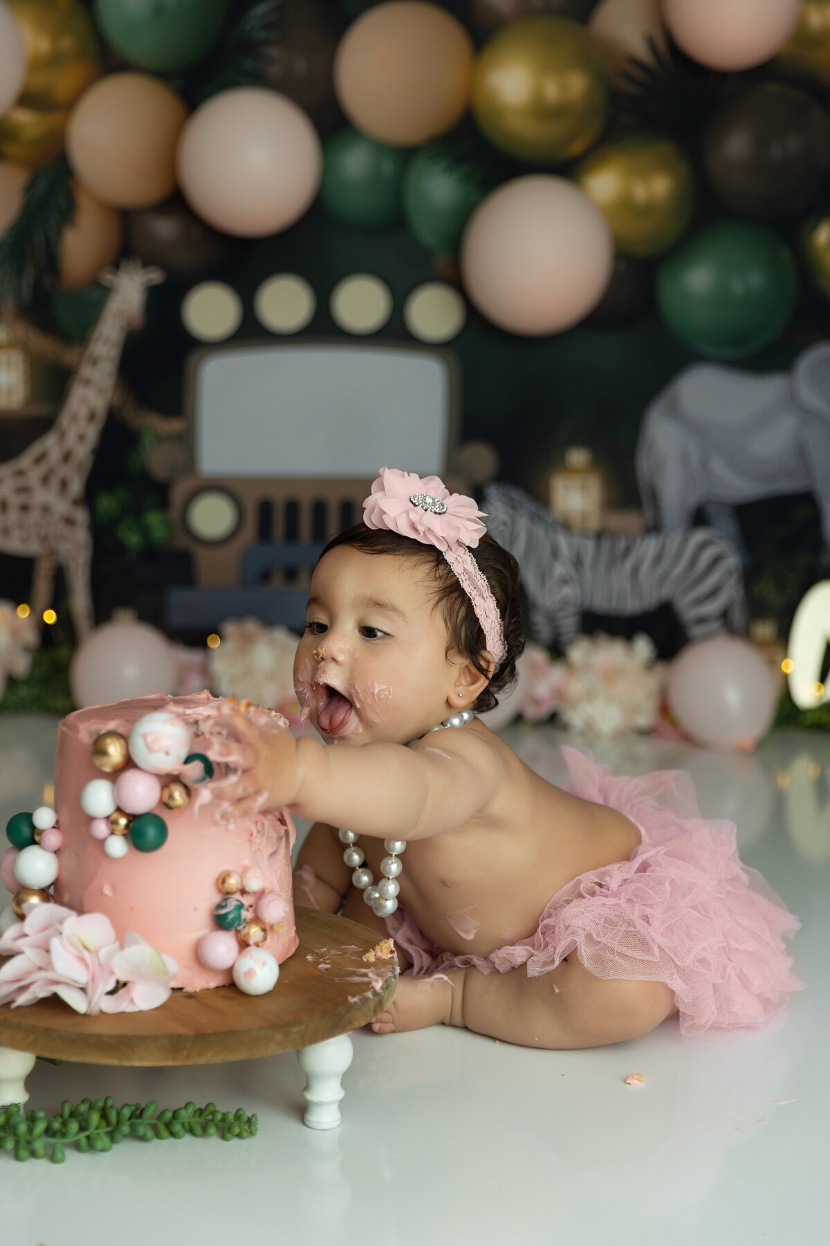A toddler girl digs into a pink cake while wearing a matching tututu and flower bow in a jungle safari themed studio