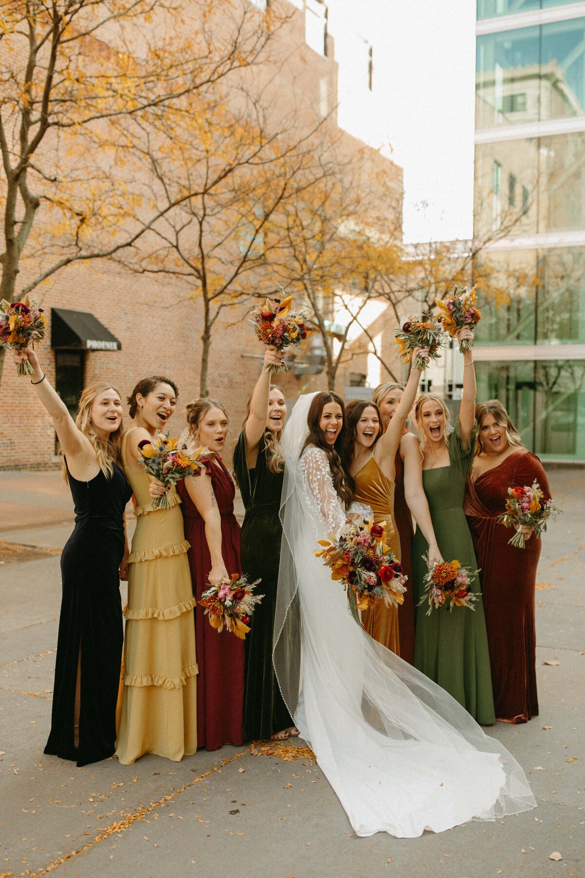 Bride and bridesmaids in colorful dresses celebrating with raised bouquets on a city street in Davenport, exhibiting joyful expressions.