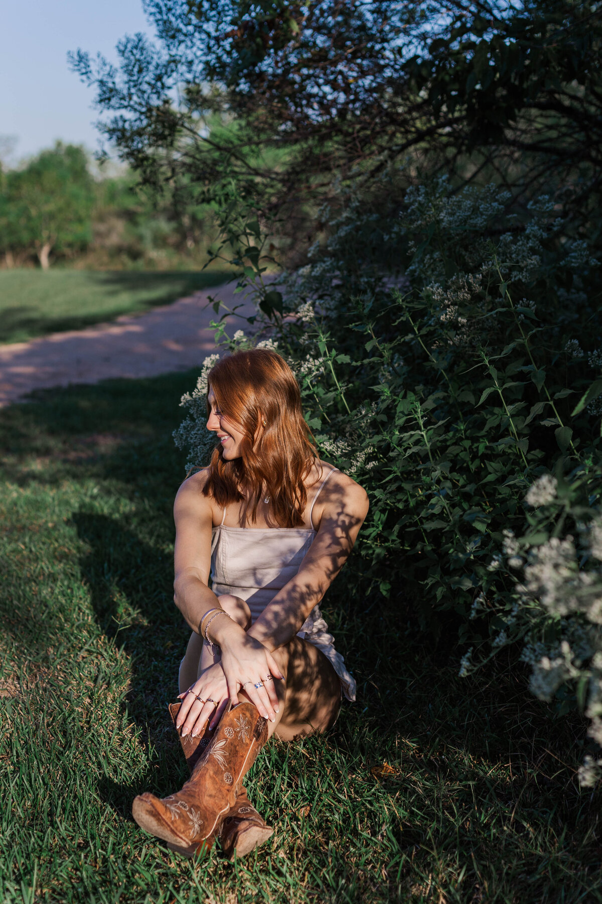 A senior girl sits in the grass while a flowery bush casts a shadow across her body