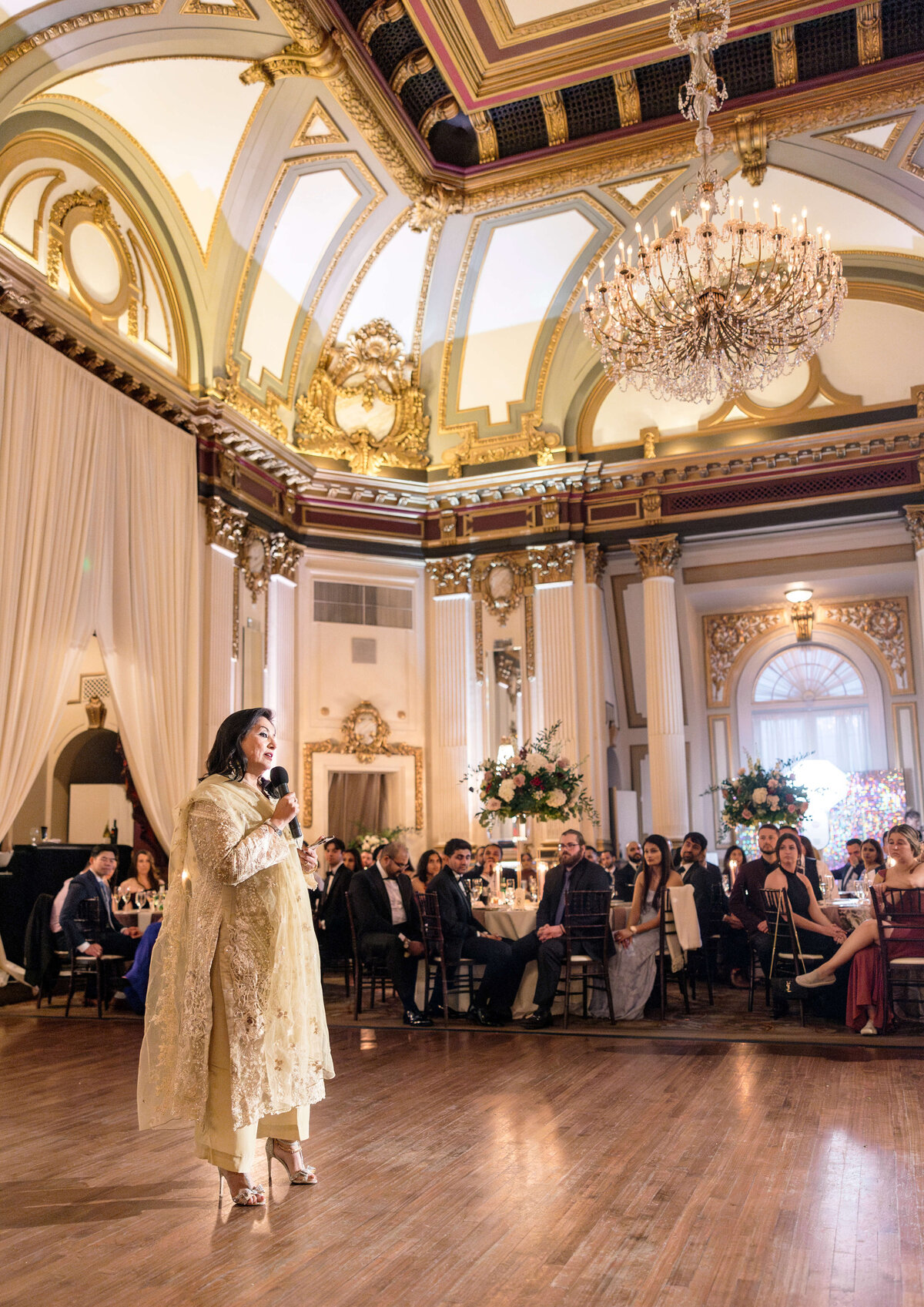 A woman in a gold dress speaks into a microphone at a grand banquet hall. Guests are seated at round tables adorned with floral centerpieces. The room features ornate architecture, chandeliers, and luxurious drapery.