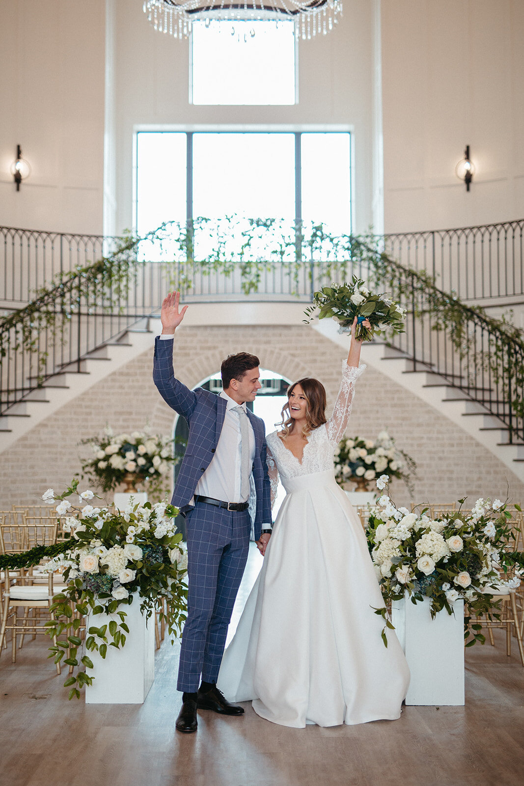 Bride and groom in a blue suit and white wedding gown wave in front a staircase wrapped in garland and white flowers