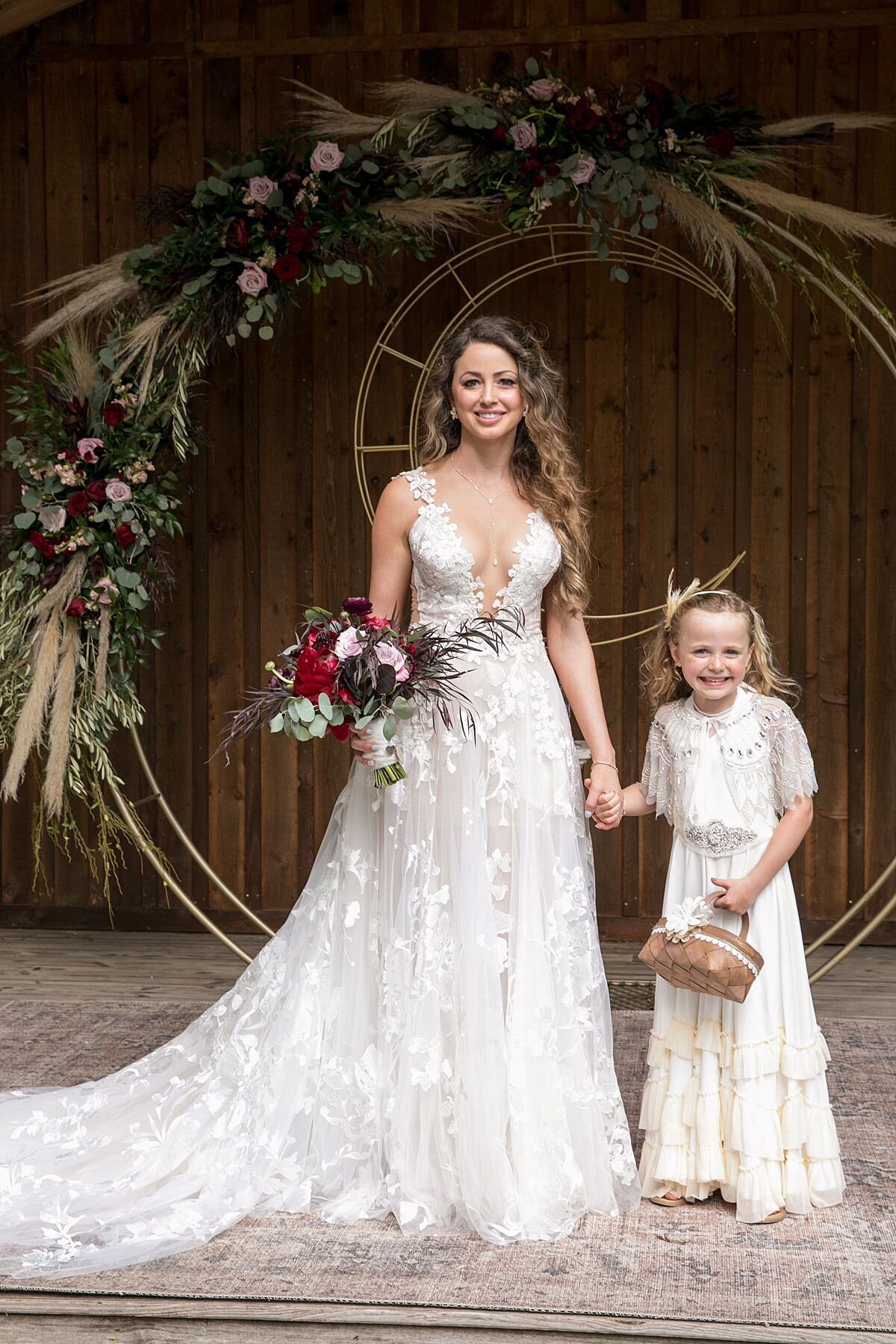 The bride, wearing a long lace wedding gown with a plunging neckline, holding her blush and burgundy bridal bouquet of peonies, ranunculus and roses holds the hand of the flower girl in the rustic log ceremony pavilion at Saddle Woods Farm. The flower girl, wearing a 1920s white lace floor length dress holds hands with the bride and she clings tightly to her light brown wicker basket. They are standing in front of the gold circular ceremony arbor with a crescent moon, decorated with pampas grass, blush and burgundy flowers and greenery.