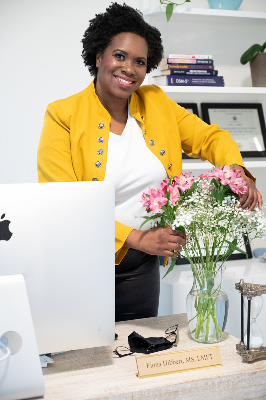 A portrait of a woman on an office. she is behind the desk fixing a flower vase and smiling to the camera