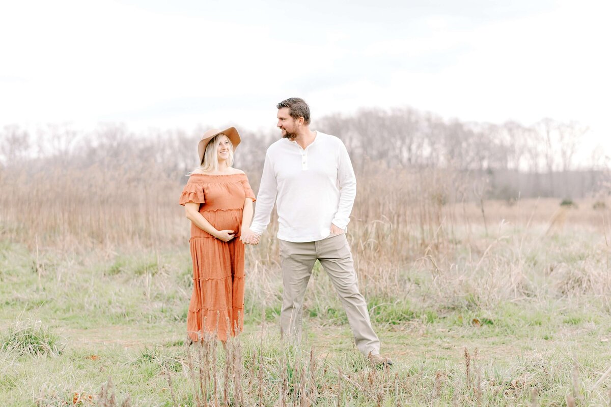 An expecting couple walk hand in hand in a field with tall golden grass during the winter while clutching her baby bump.