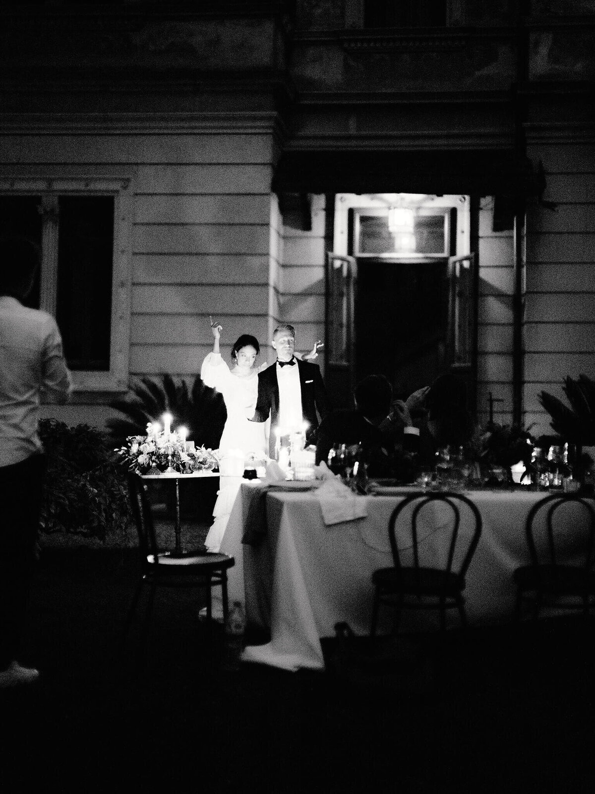 Bride and groom standing, the bride's right arm is raised in the air, with a dining table in front and a villa at the back.