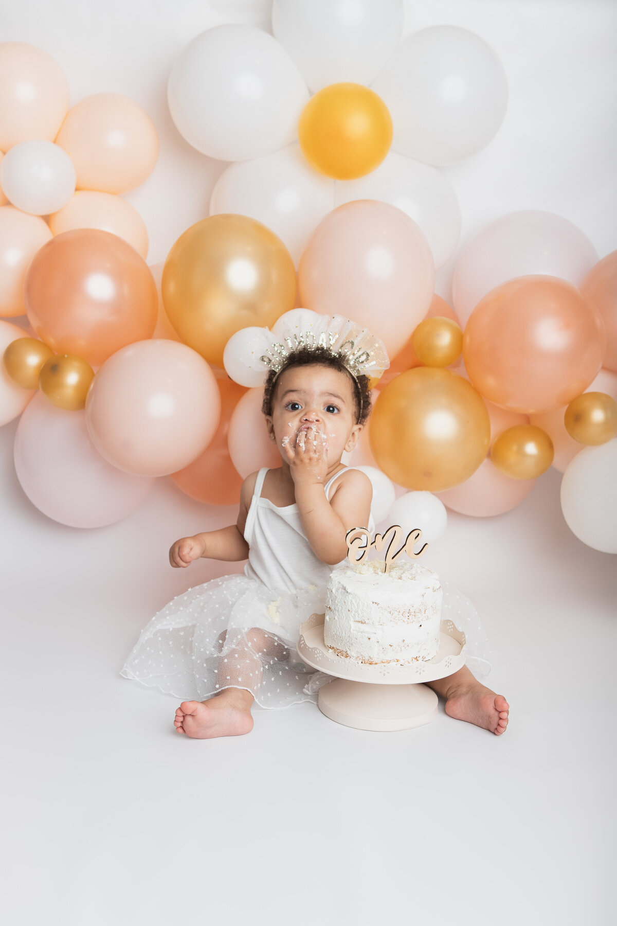 Baby eating a messy white cake with a peach balloon garland