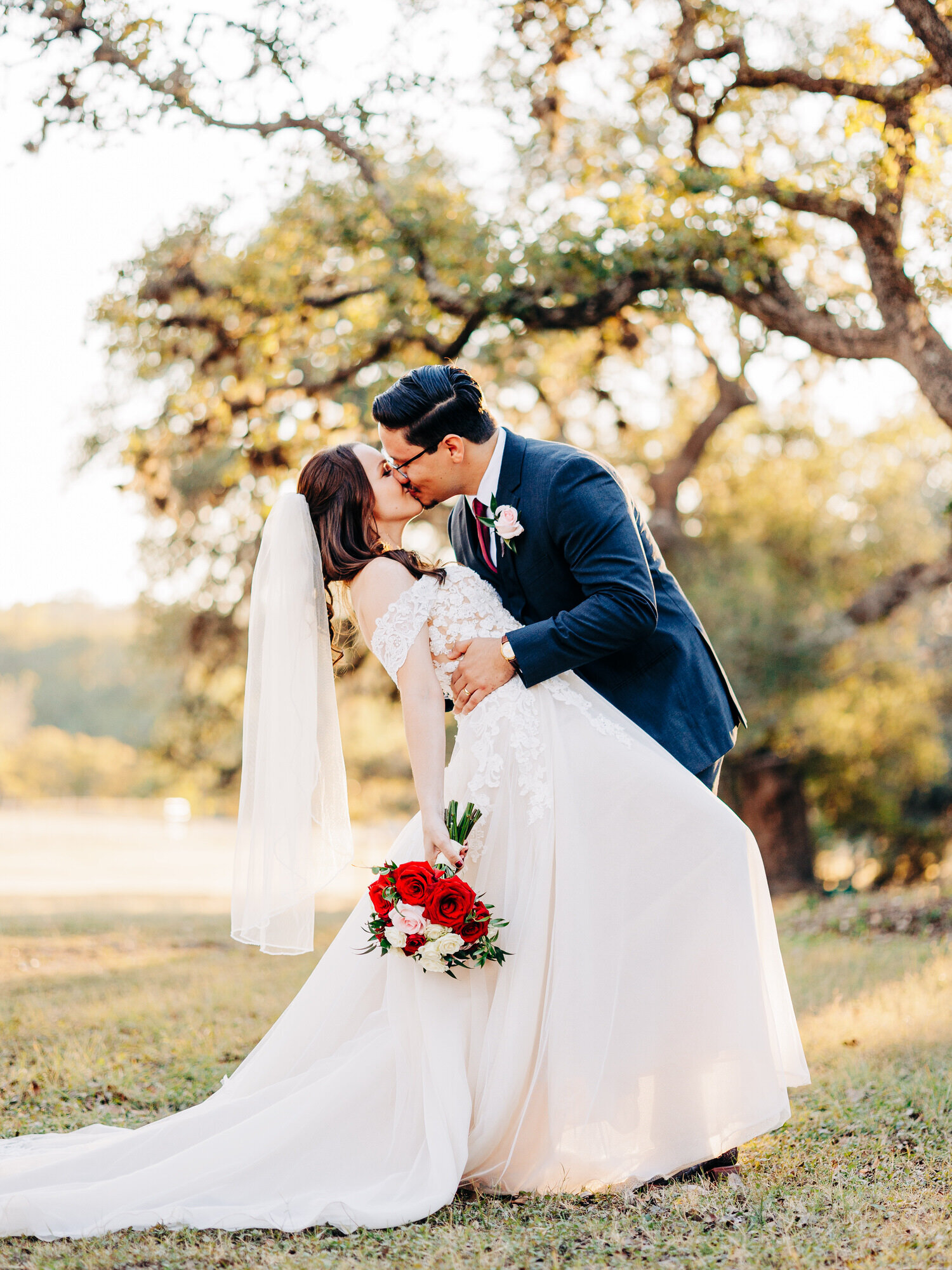 This image features a bride and a groom at a fall wedding. The groom, wearing a blue suit and a burgundy tie,  is dipping his bride while kissing her. The bride is wearing a mid-length veil. The image was taken by KD Captures, a wedding photographer in San Antonio.