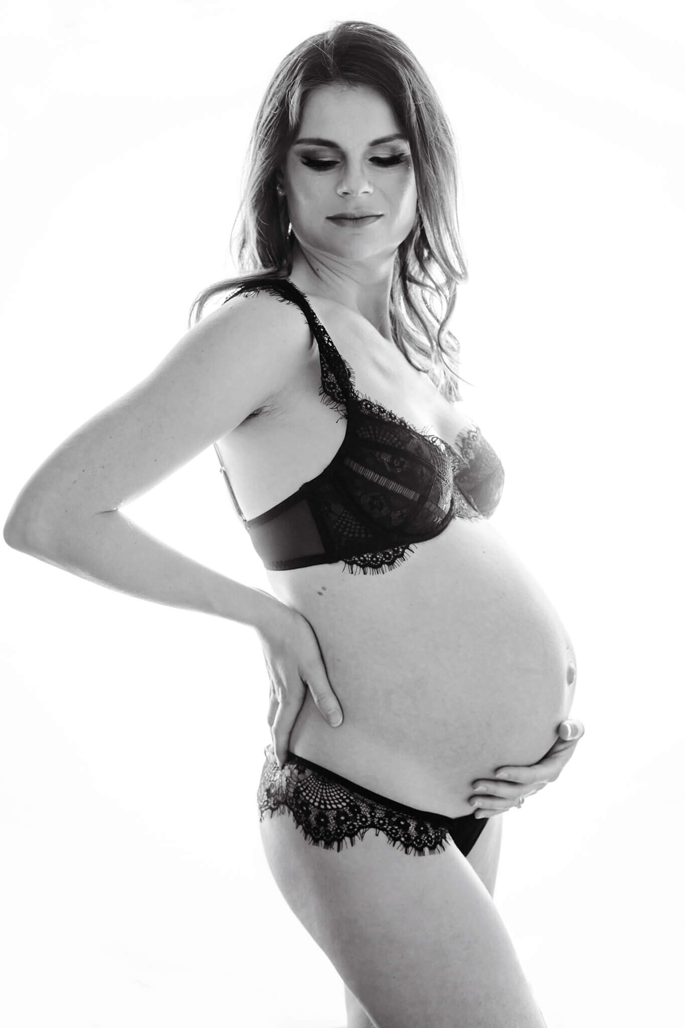 pregnant woman is looking down, one hand on her waist and the other one on her belly, she is wearing black lingerie for her maternity photoshoot