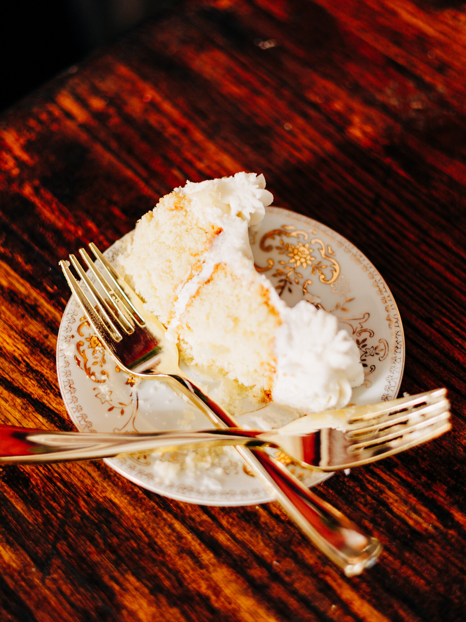 This image features a wedding cake on a vintage plate that has been cut by the bride and groom. Two gold forks are sitting on the plate, crossing over each other. The cake is white with white frosting.