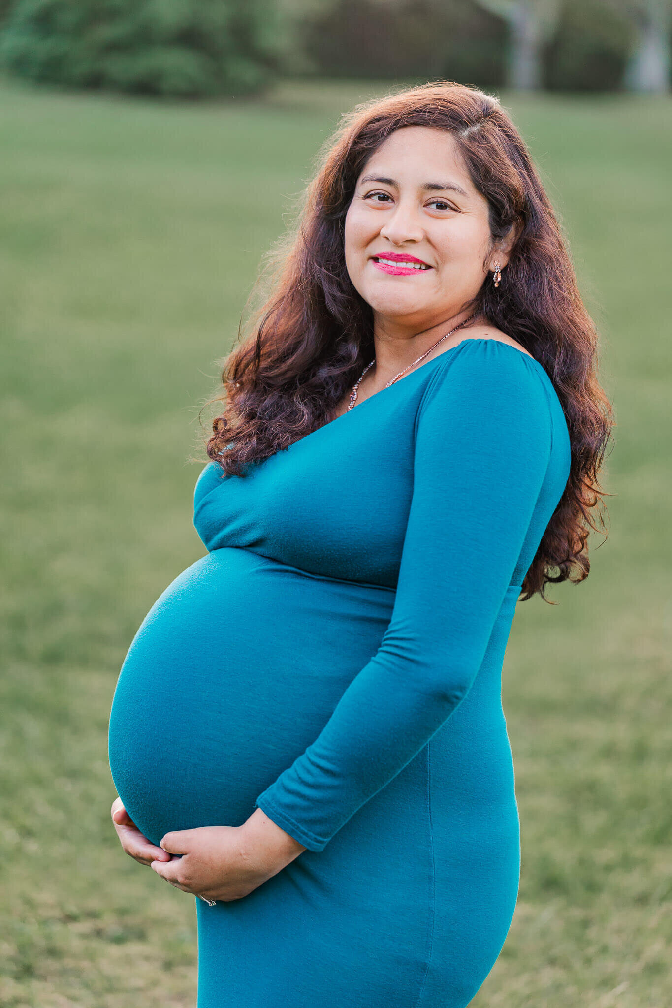A pregnant woman in blue hugging her belly at a park in Burke at sunset.
