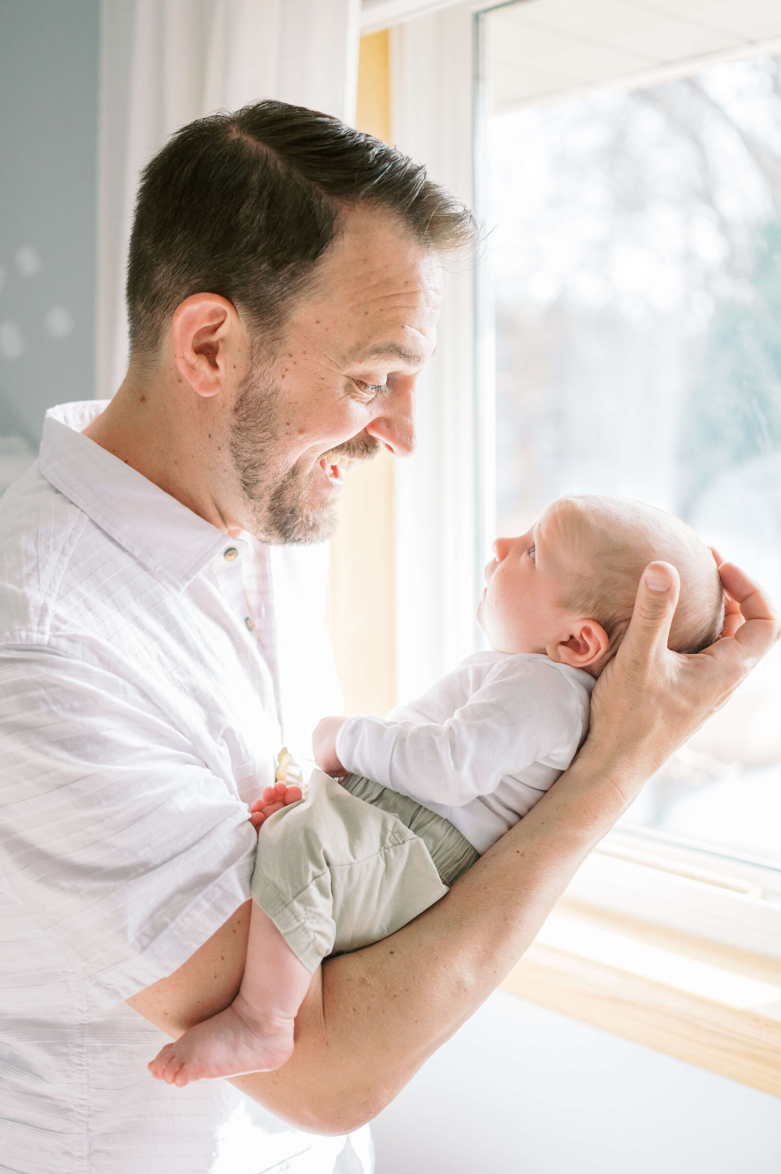 A dad, dressed in a white collared shirt, holds his infant son and gives him a big smile