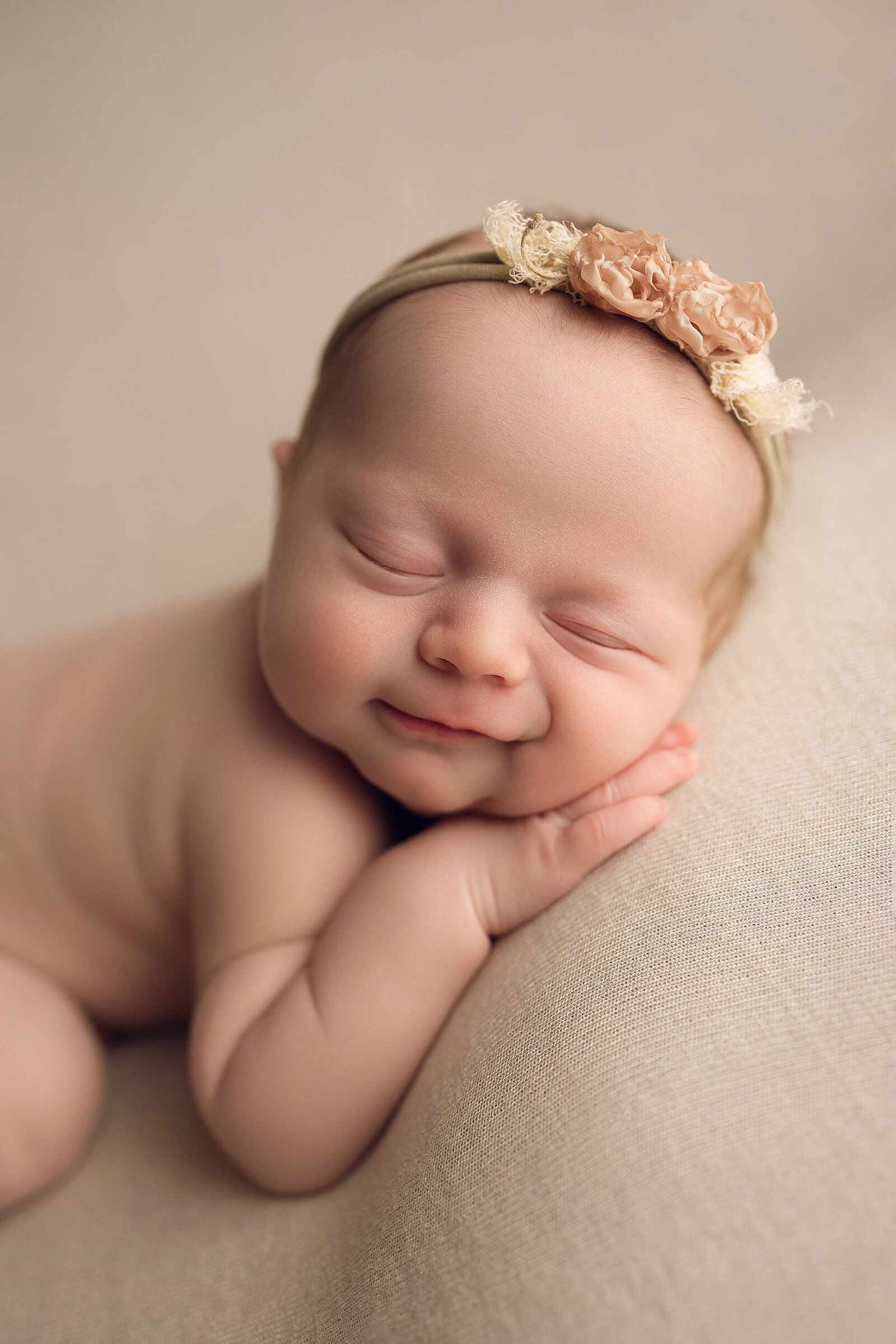 A Caucasian baby lays on a tan blanket and smiles with her eyes closed.