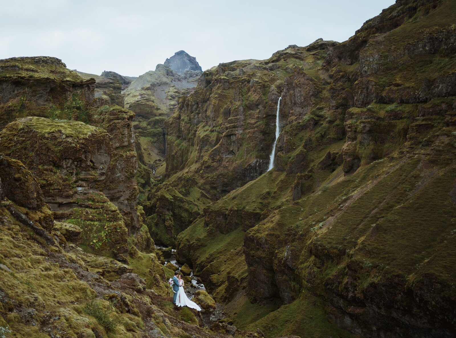 bride and groom standing in front of the mountains and canyon with a waterfall running behind them. They are embracing each other from far away.