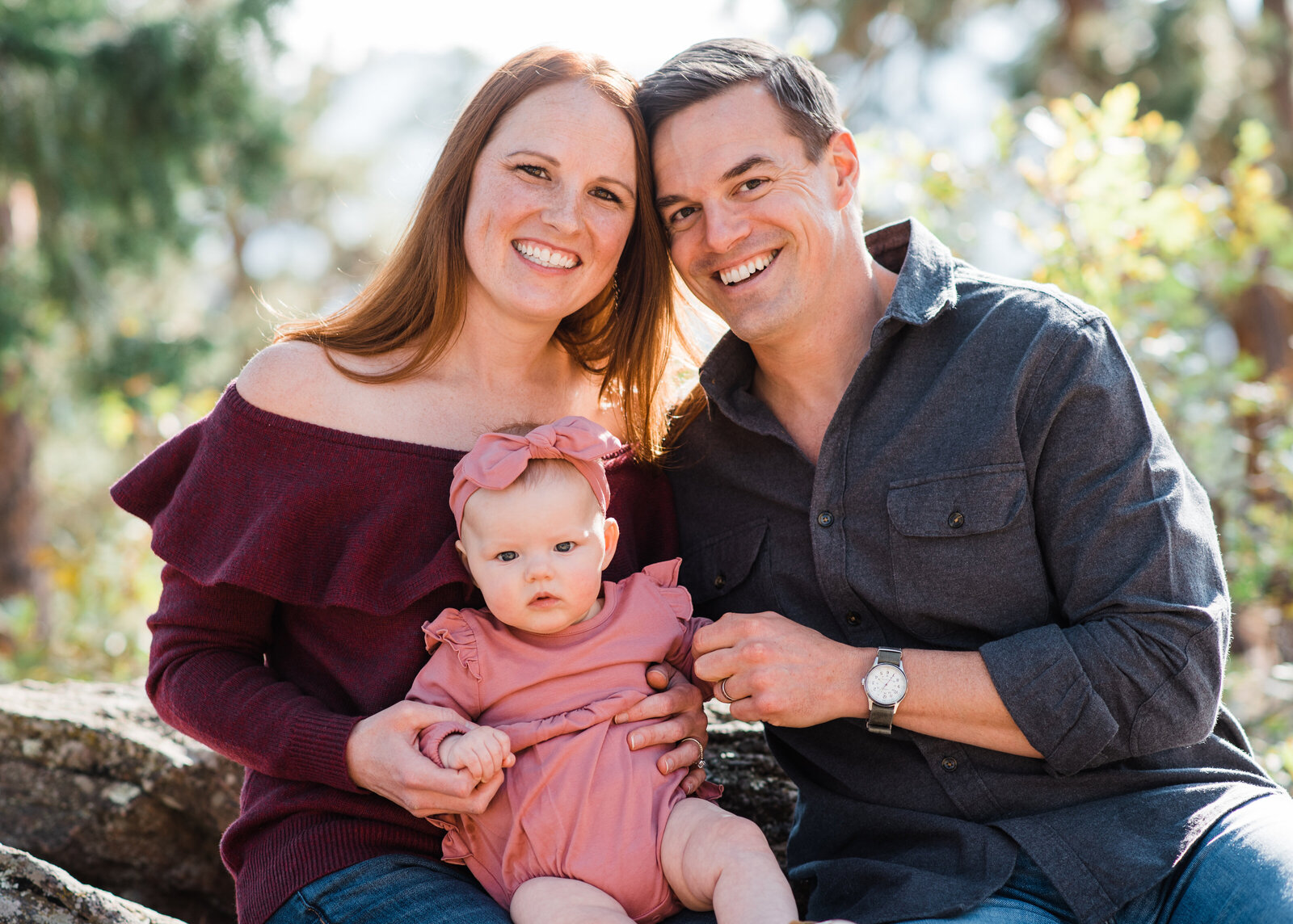 denver family photographers captures young family with their baby girl sitting togethre in the woods of the rockies leaning into each other and cuddling close for their family outdoor photos