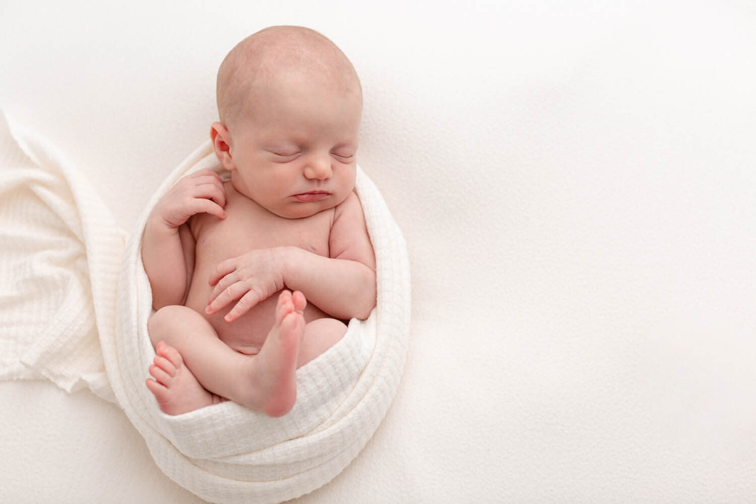 Newborn baby swaddled in white and sleeping peacefully on a white backdrop.