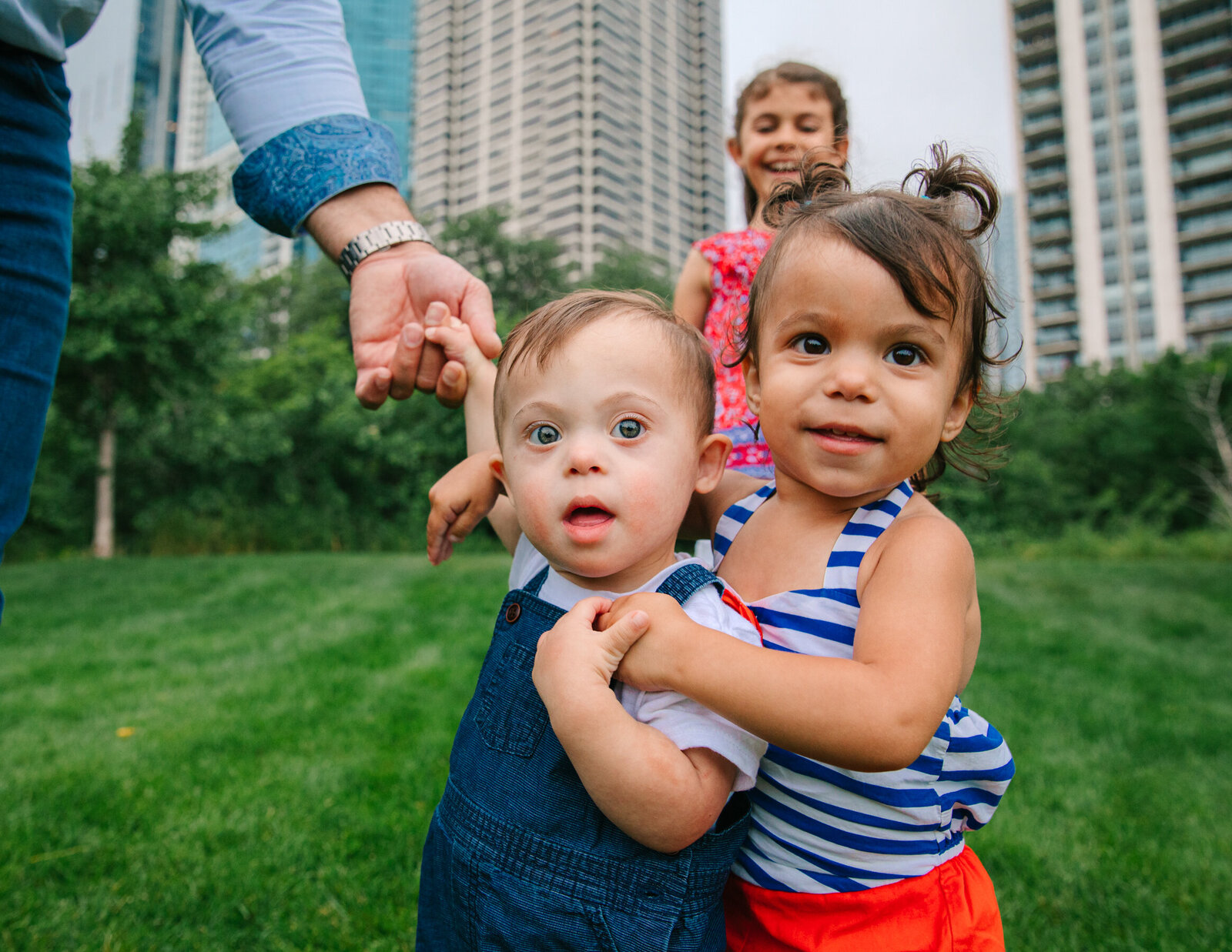 photo of one year old twins, a girl  and a boy with down's syndrome, on green grass with older sister in the background