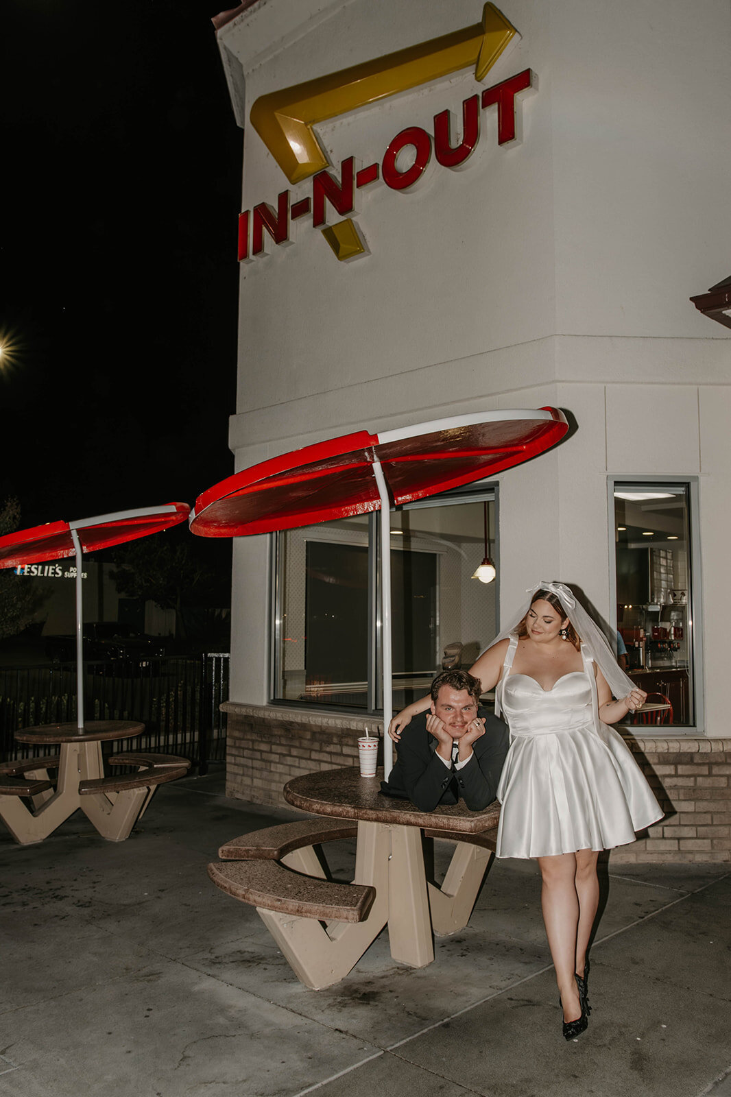 A bride and groom pose outside an In-N-Out Burger at night, sitting and standing by a picnic table, captured in candid wedding photography at golden hour.