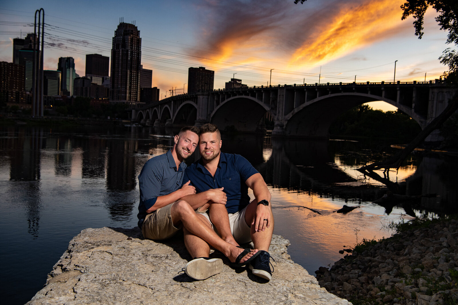 Gay couple smile while snuggling on a rock at sunset in Minneapolis, Minnesota.