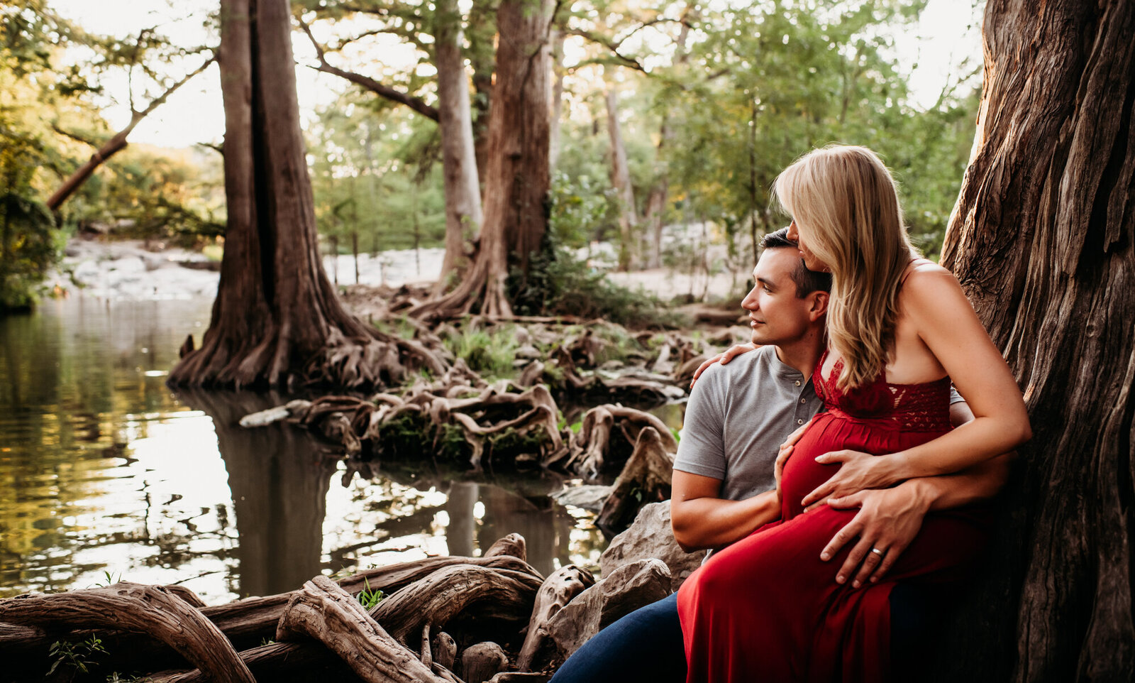 Maternity Photographer, Pregnant woman in a red dress leaning against a tree while she is sitting on a man's lap in a gray t shirt. They are next to a creek and Cypress trees.