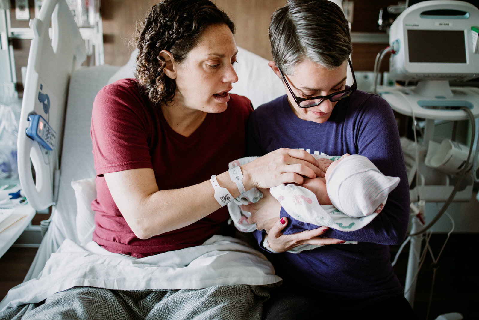two moms in the hospital sitting on hospital bed with newborn baby girl
