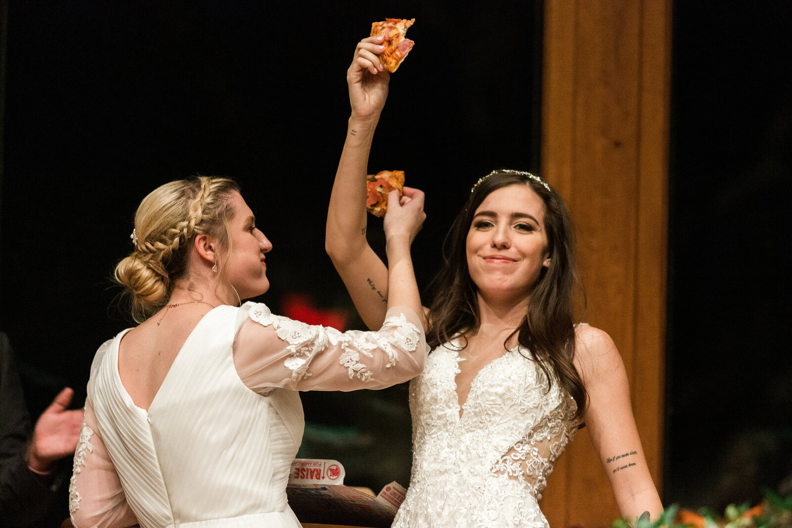 Two brides sharing "unity pizza" during their wedding ceremony. The bride on the left is wearing a mid-sleeve white dress while the bride on the right is wearing a sleeveless white dress. Both are holding up their respective slices of pizza joyfully in the air.