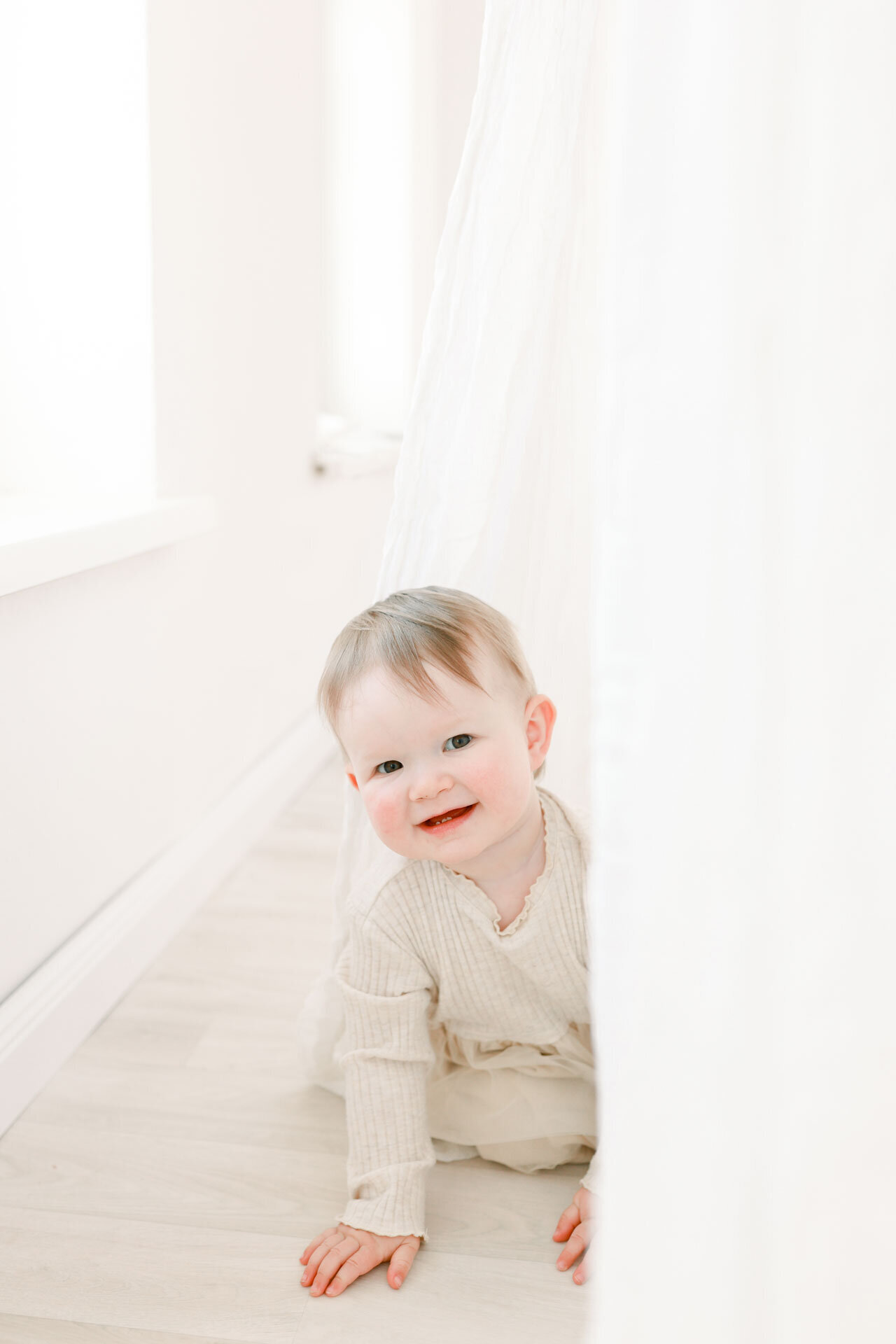 Ein verspieltes Baby, das lächelnd hinter einer weißen Tüllgardine hervorsieht. Entstanden bei einem Babyfotoshooting bei Babyfotografin Bielefeld.
