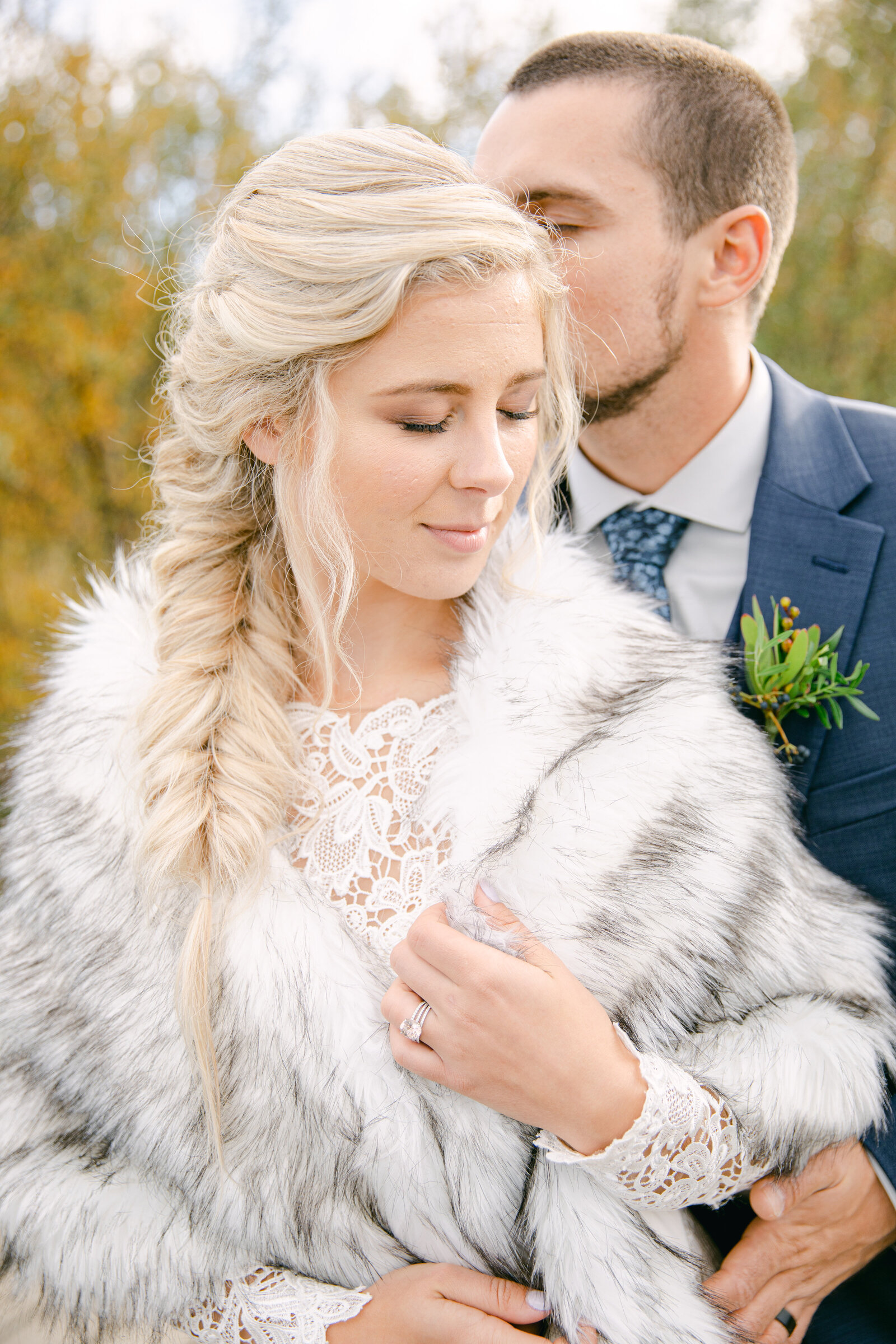 bride looking at the ground holding her fur coat as groom kisses the side of her head