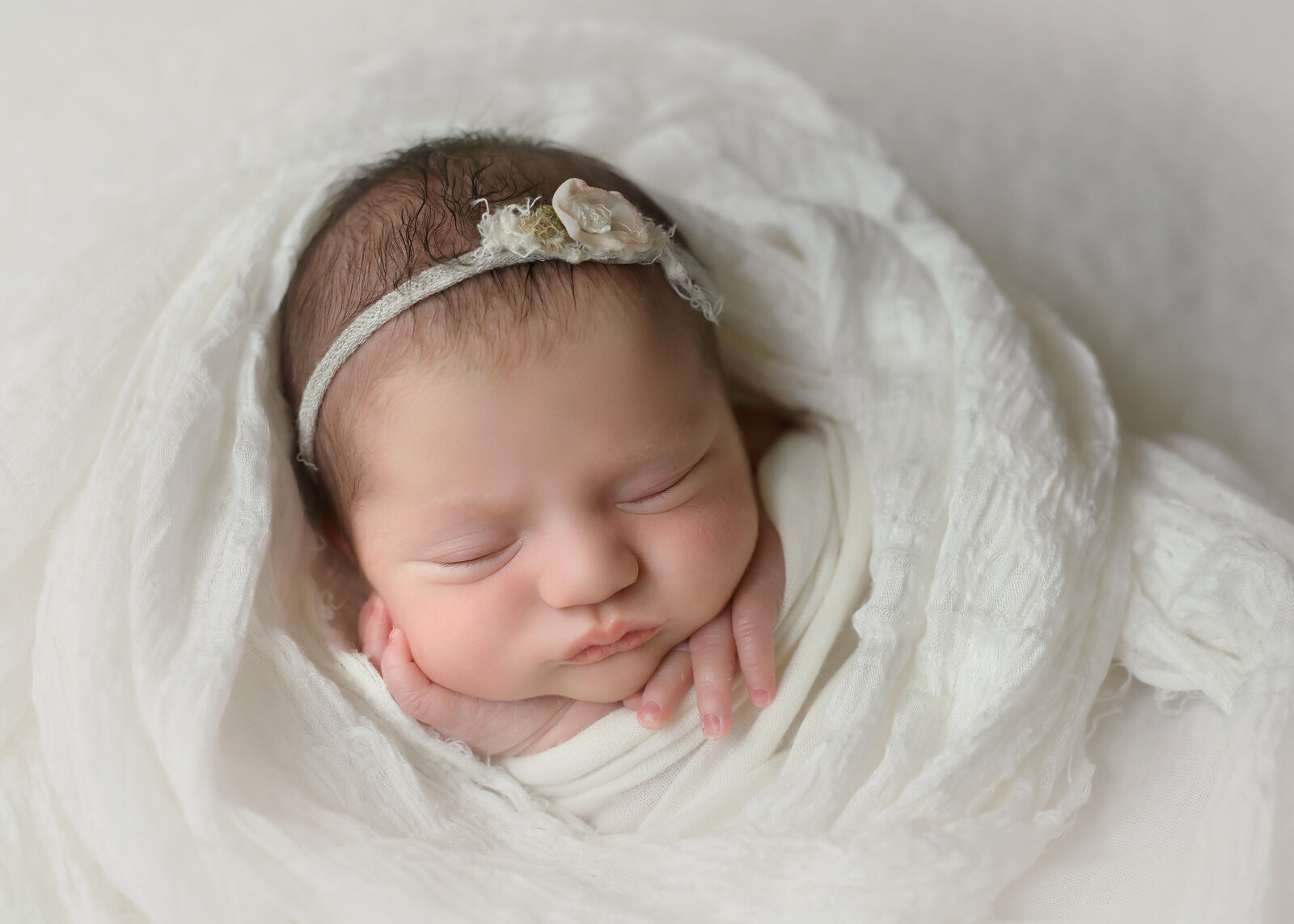 newborn baby girl with headband sleeping and swaddled in cream blanket