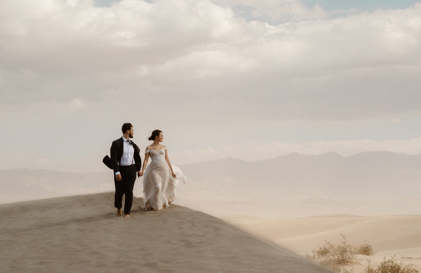 Barefoot bride and groom walking hand in hand across a sand dune in Death Valley, with distant mountains and dramatic clouds in the background.