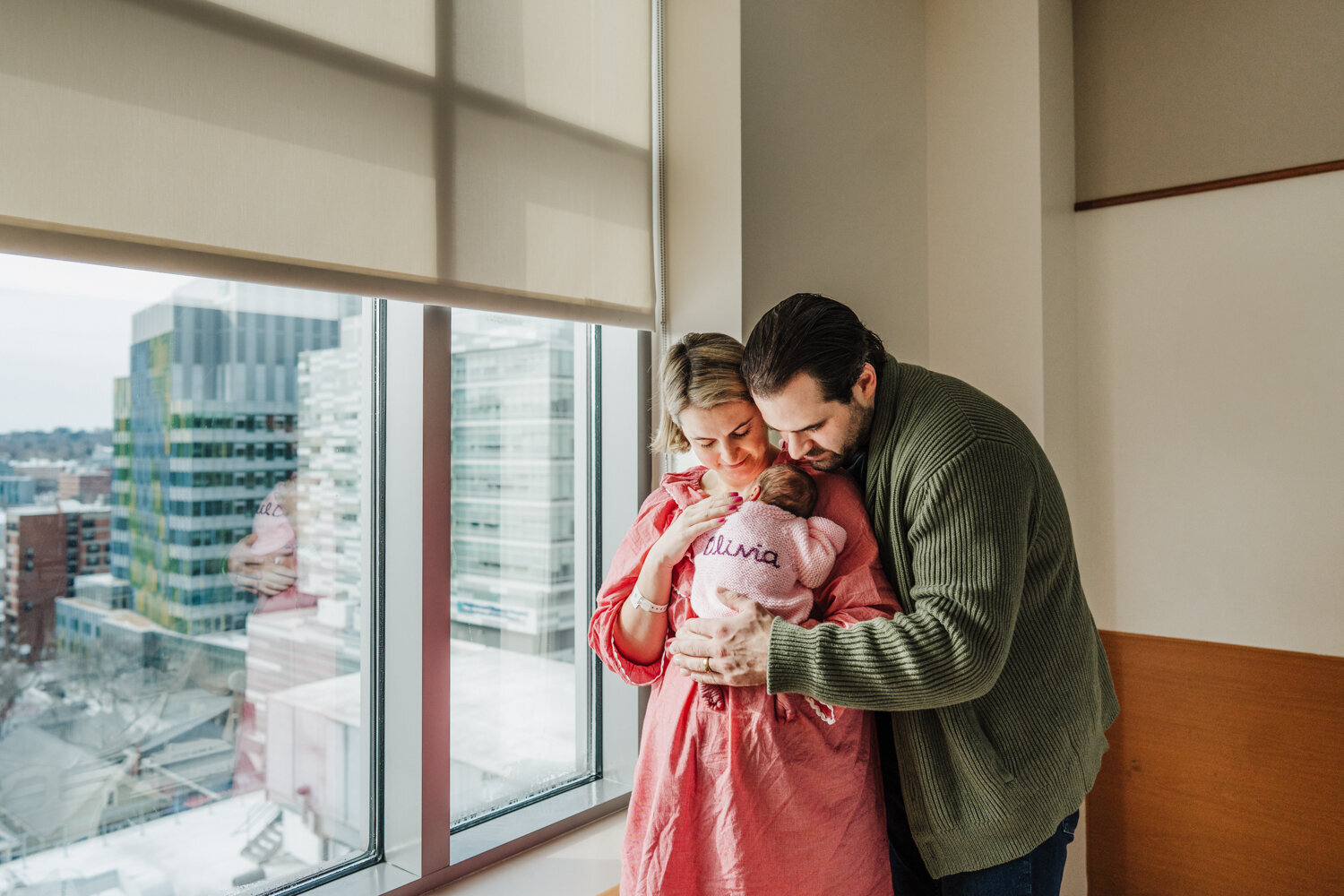 parents smile down at new baby girl in front of hospital window