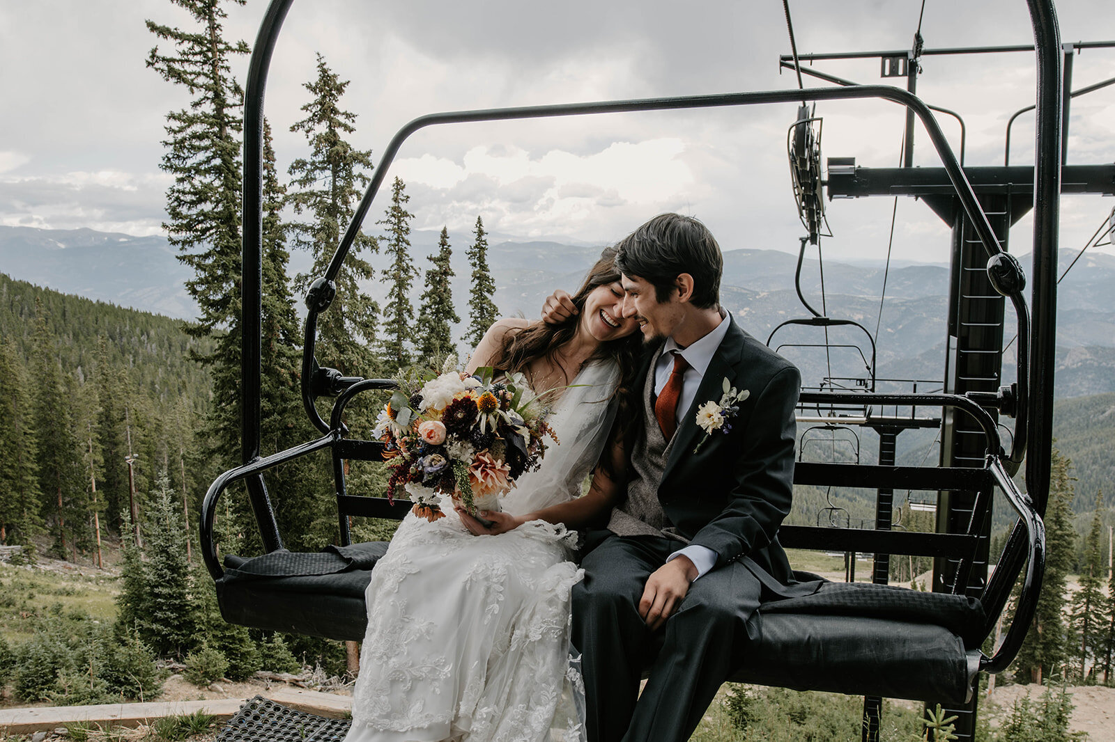Bride and groom sitting closely on a ski lift during their Colorado wildflower elopement, surrounded by mountains and pine trees, smiling happily as their real connection is captured.