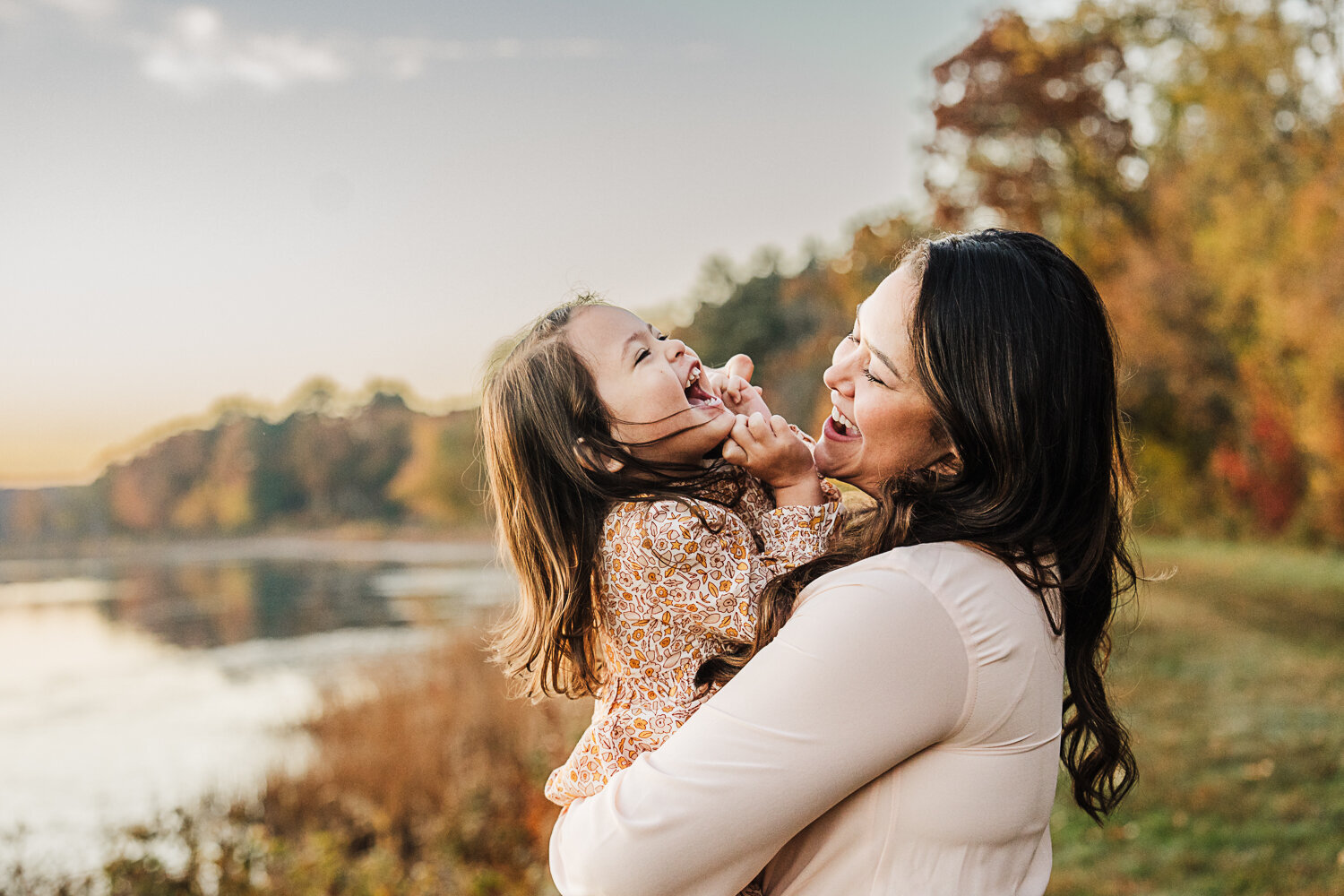 mom holds toddler daughter and both laugh beside a lake at sunrise
