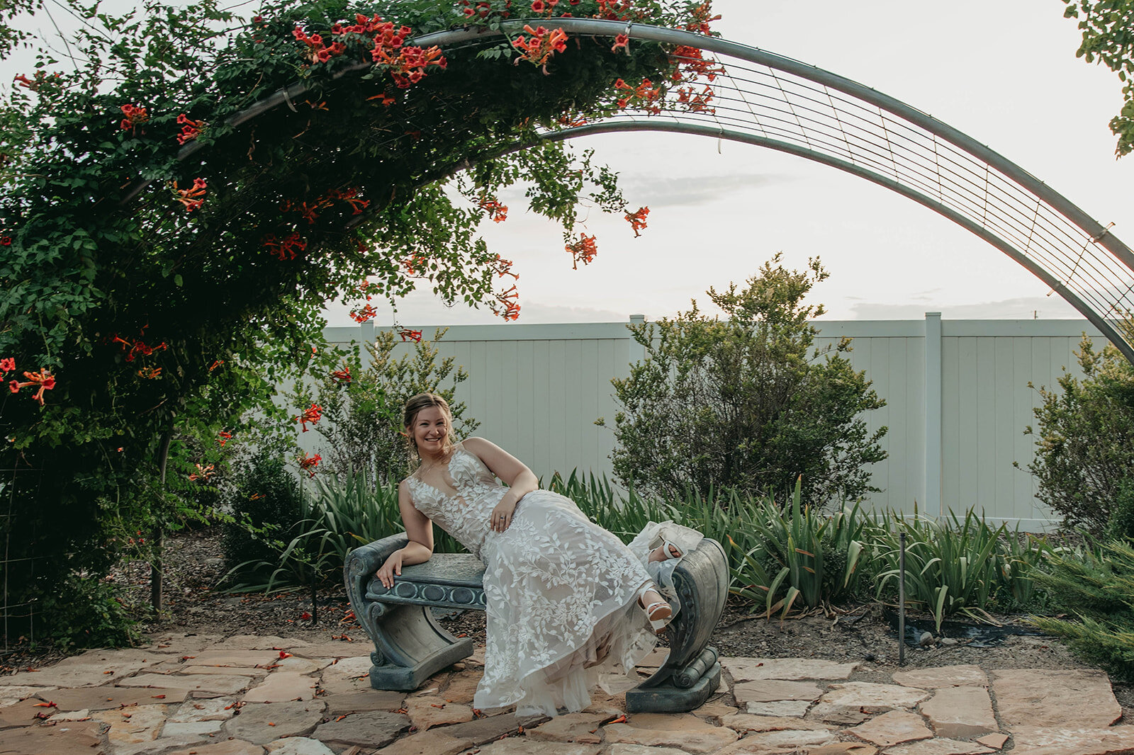 A bride in a white lace dress sits playfully on a sculpted bench under a flower-covered arch, capturing the charm of an intimate wedding in the Colorado mountains.