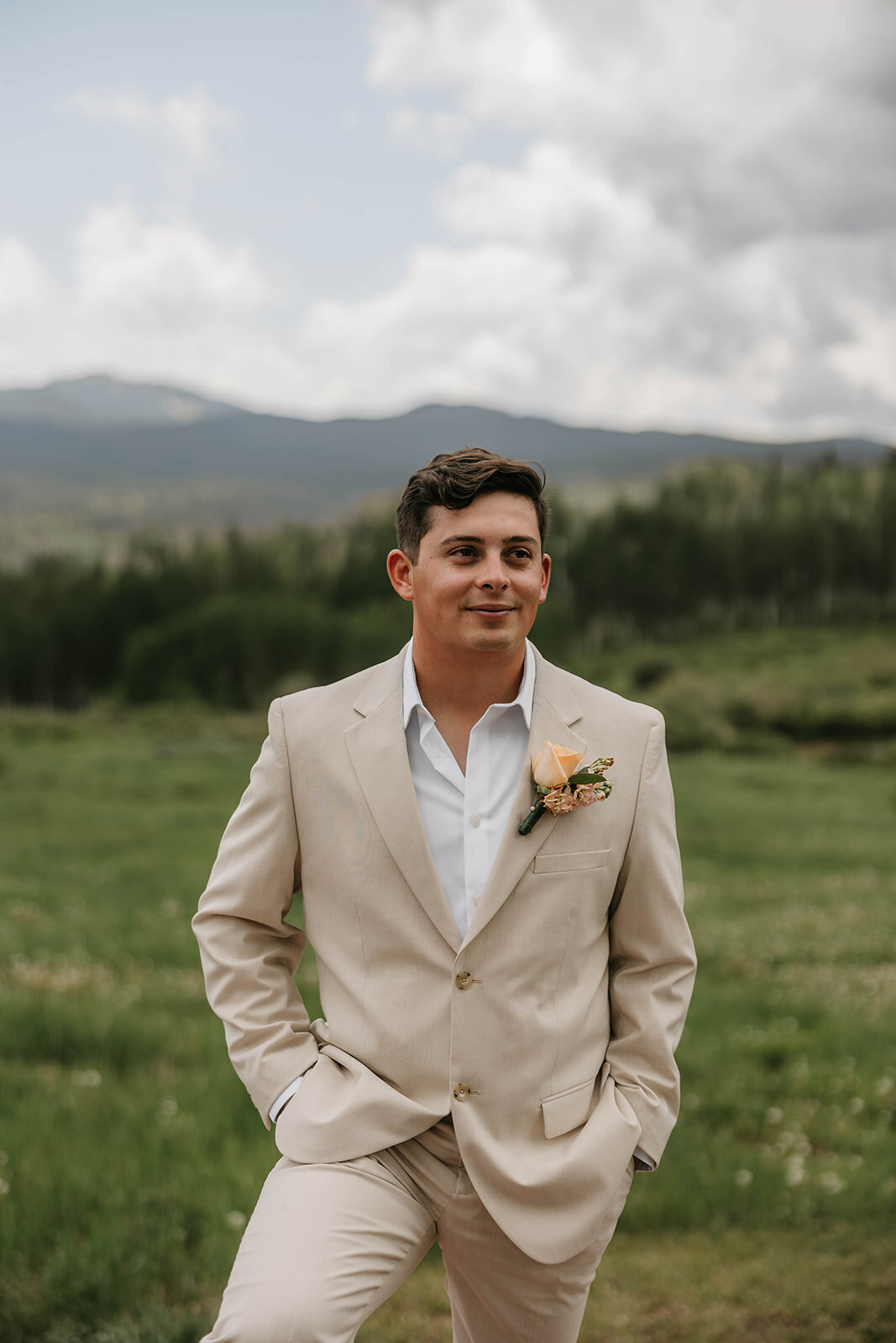 Man in a beige suit with a boutonniere stands outside in a green field with mountains in the background, capturing the spirit of an intimate wedding in the Colorado mountains.